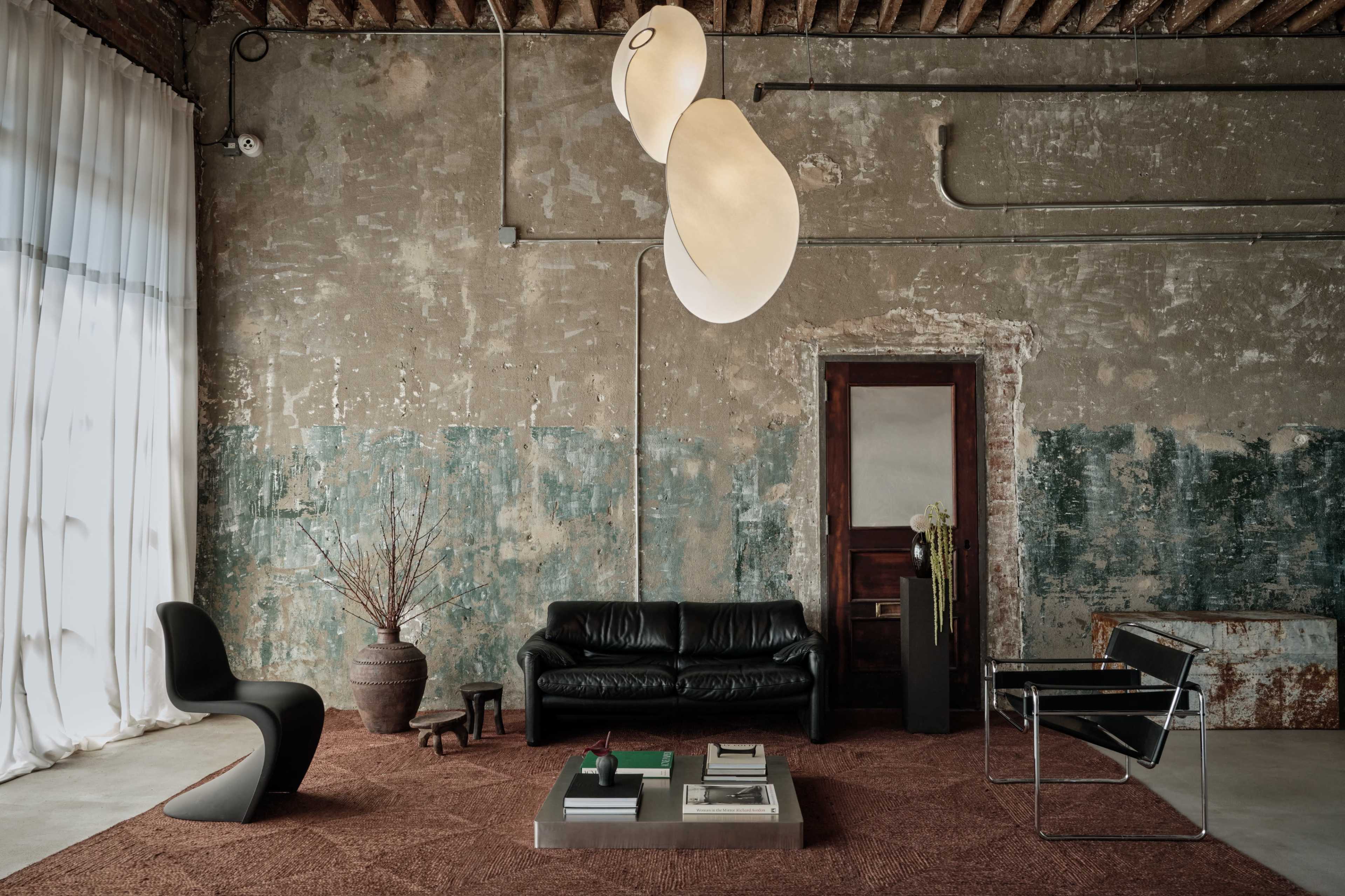 The image shows a minimalist living space with a black leather sofa, a glass coffee table, and an artistic light fixture hanging from the ceiling, set against a textured wall with peeling paint.