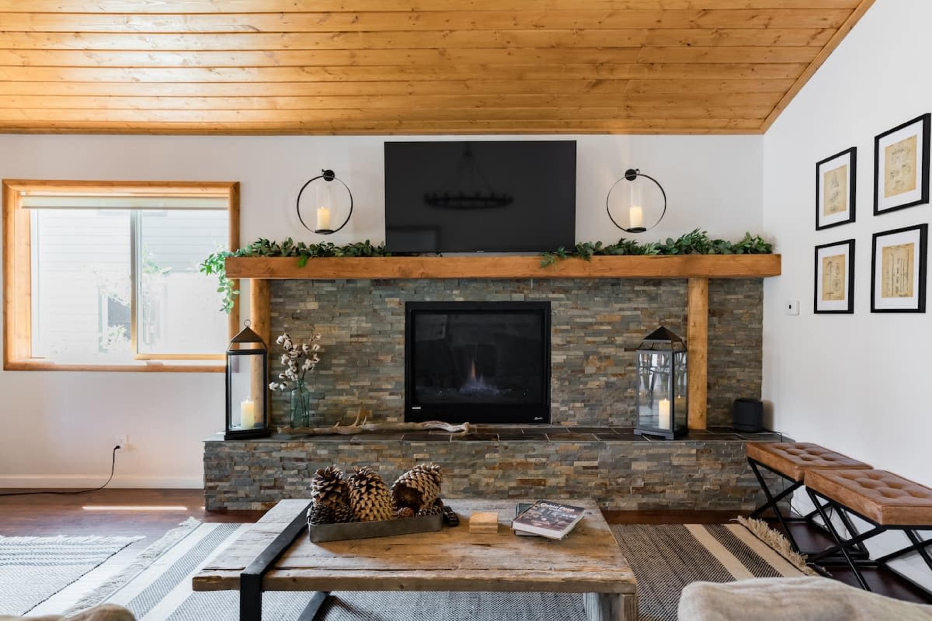 A modern living room features a stone fireplace with a wooden mantel, a television mounted above, and large windows allowing natural light to enter.