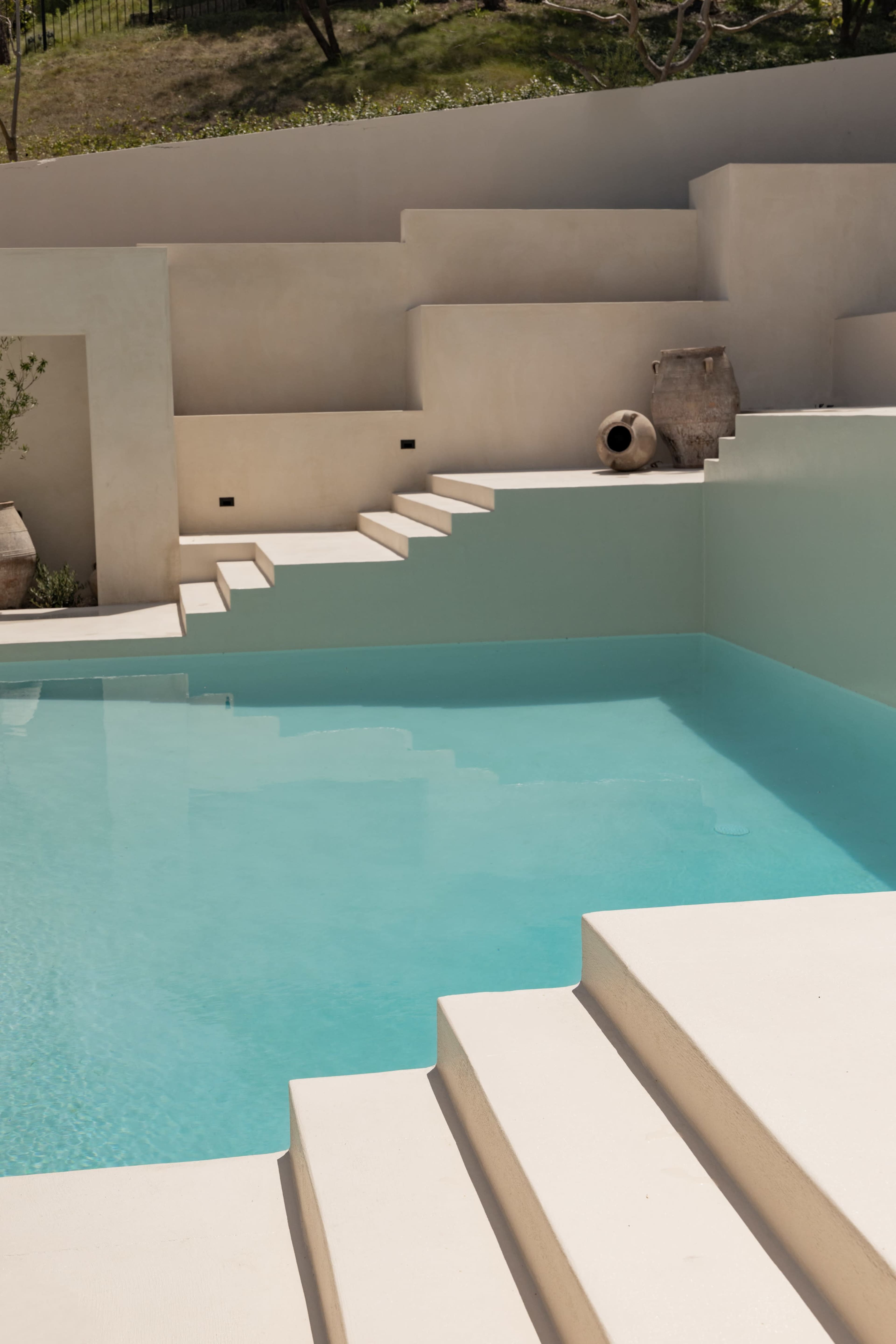 The image shows a modern swimming pool with clear turquoise water and white steps leading up to a minimalistic, textured wall.