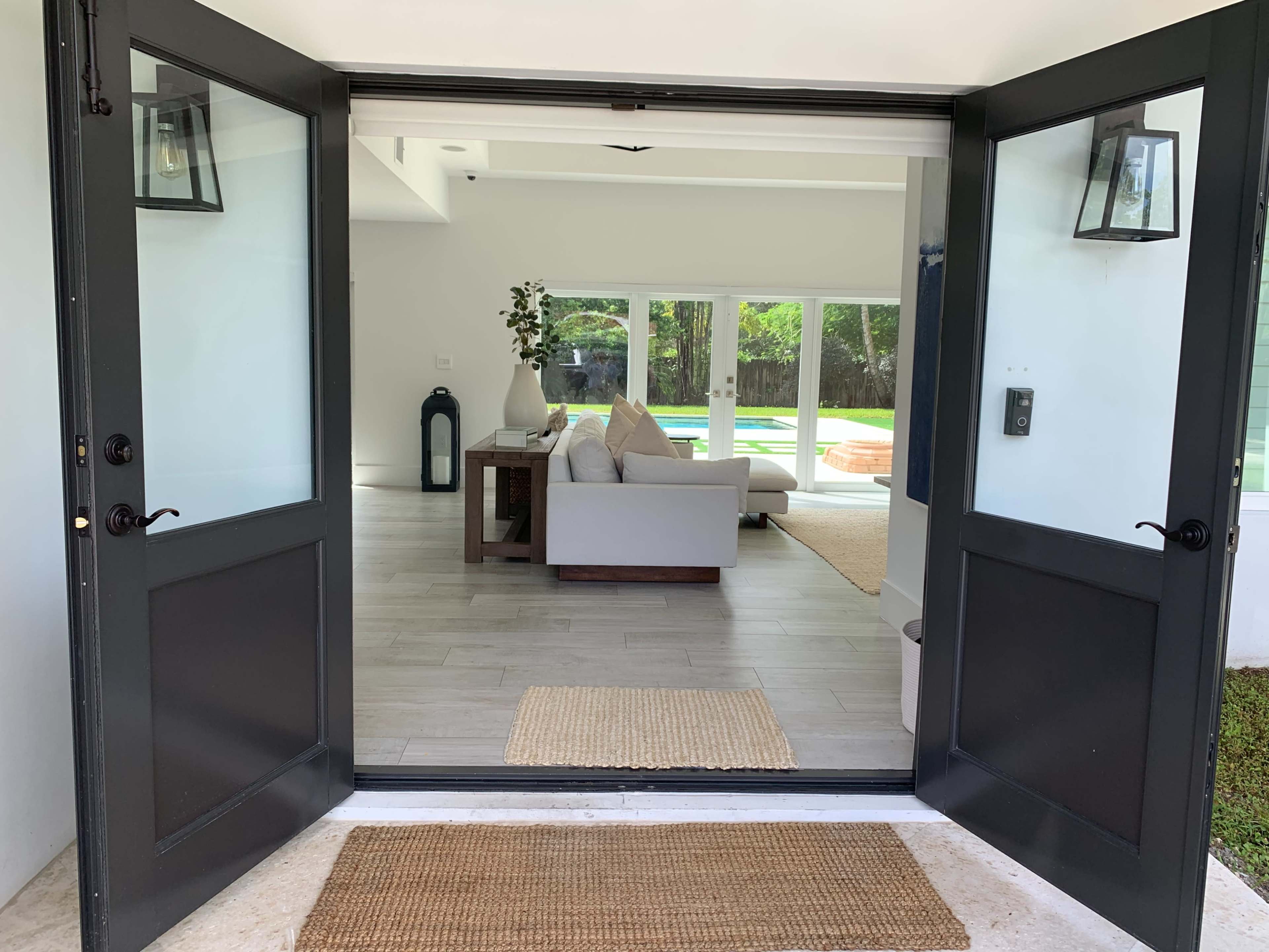 The image shows an open double door leading into a bright living space with a sofa and large windows that overlook an outdoor area.