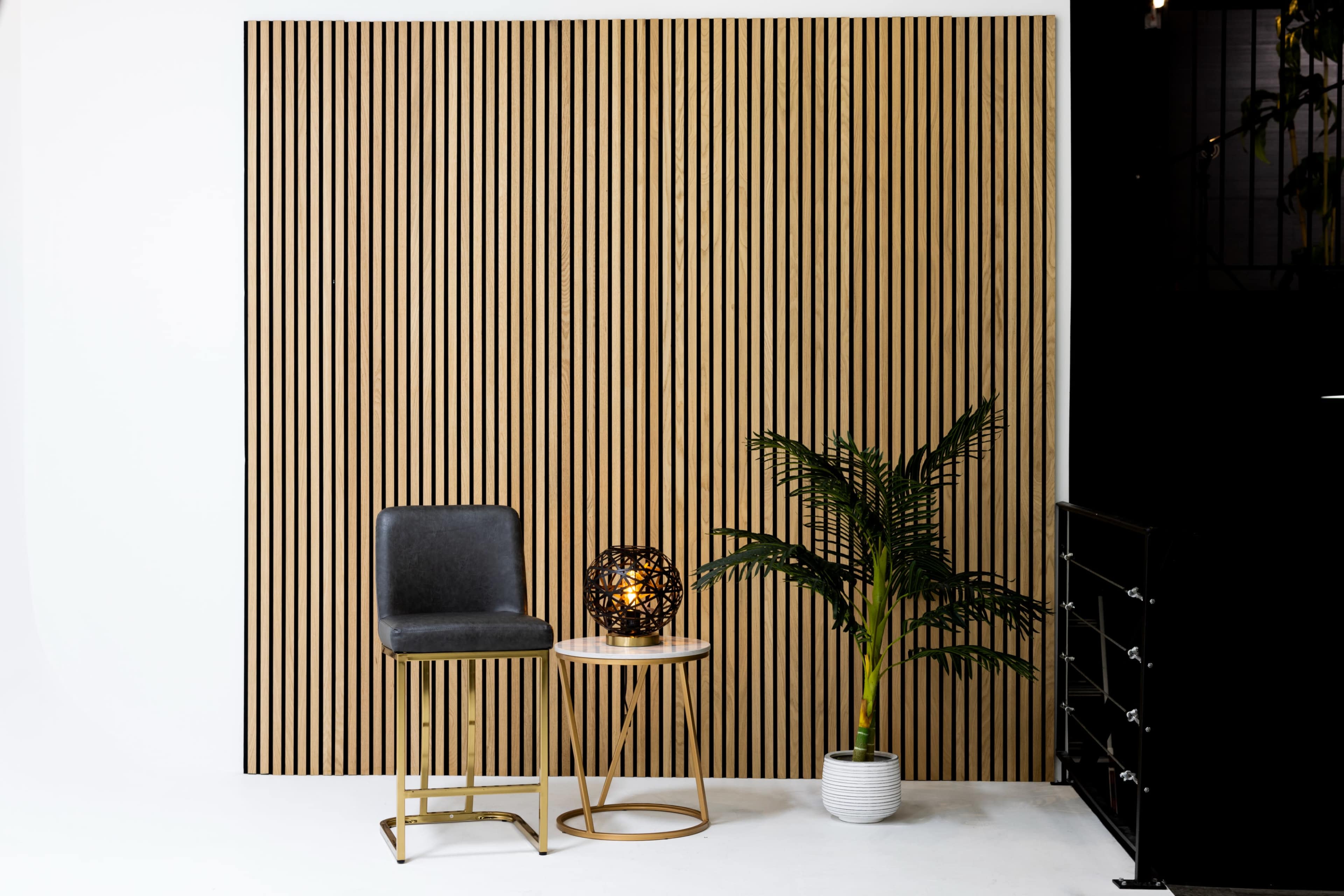 A modern interior scene with a vertical striped wooden wall, a gray stool, a round table, a decorative lamp, and a potted plant.
