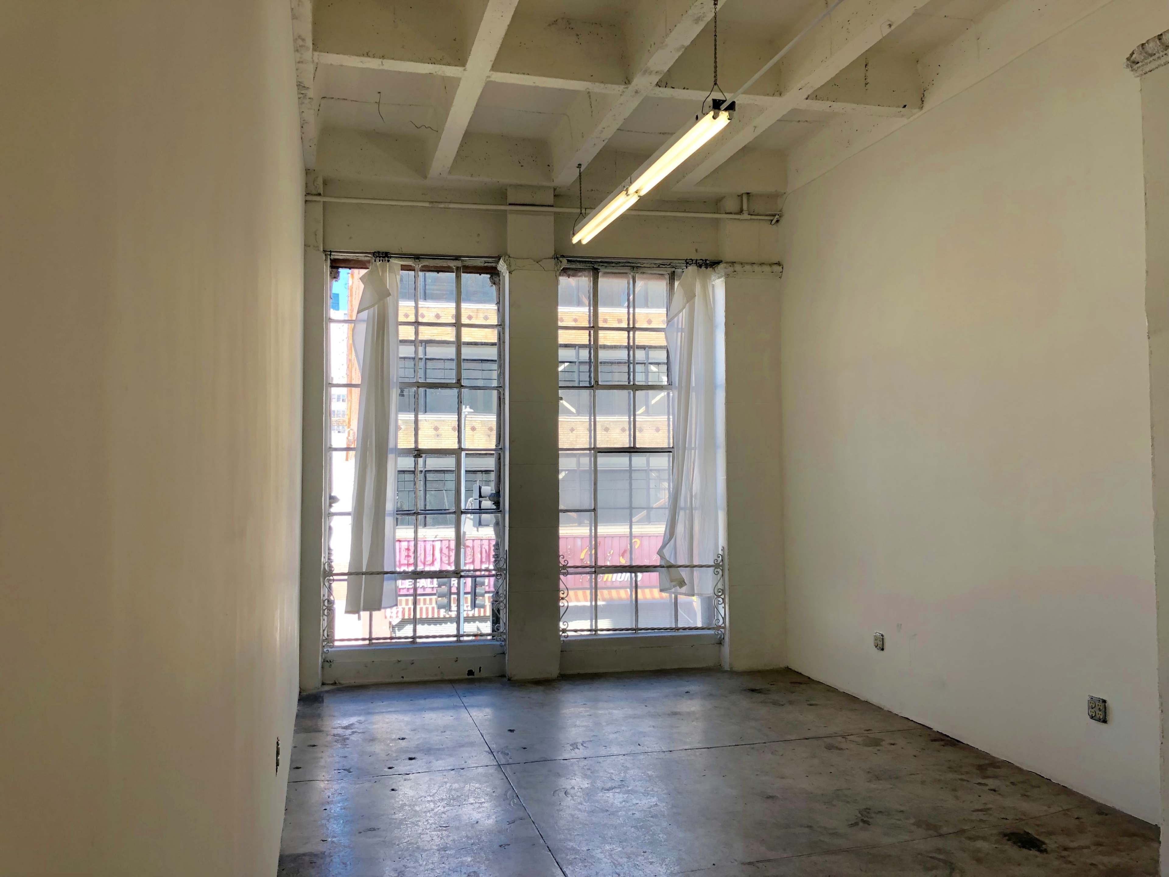 The image shows an empty room with two large windows covered by sheer curtains, featuring concrete flooring and exposed beams in the ceiling.