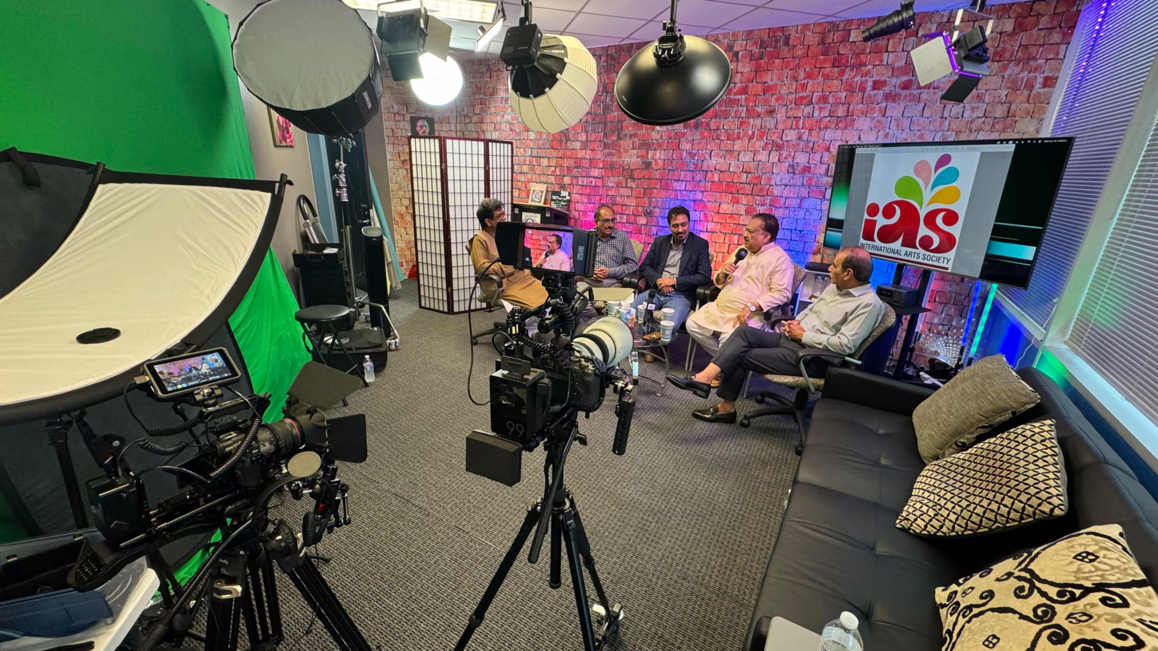 A group of five individuals is seated in a studio setting with multiple cameras and lights, discussing in front of a screen displaying a colorful logo.