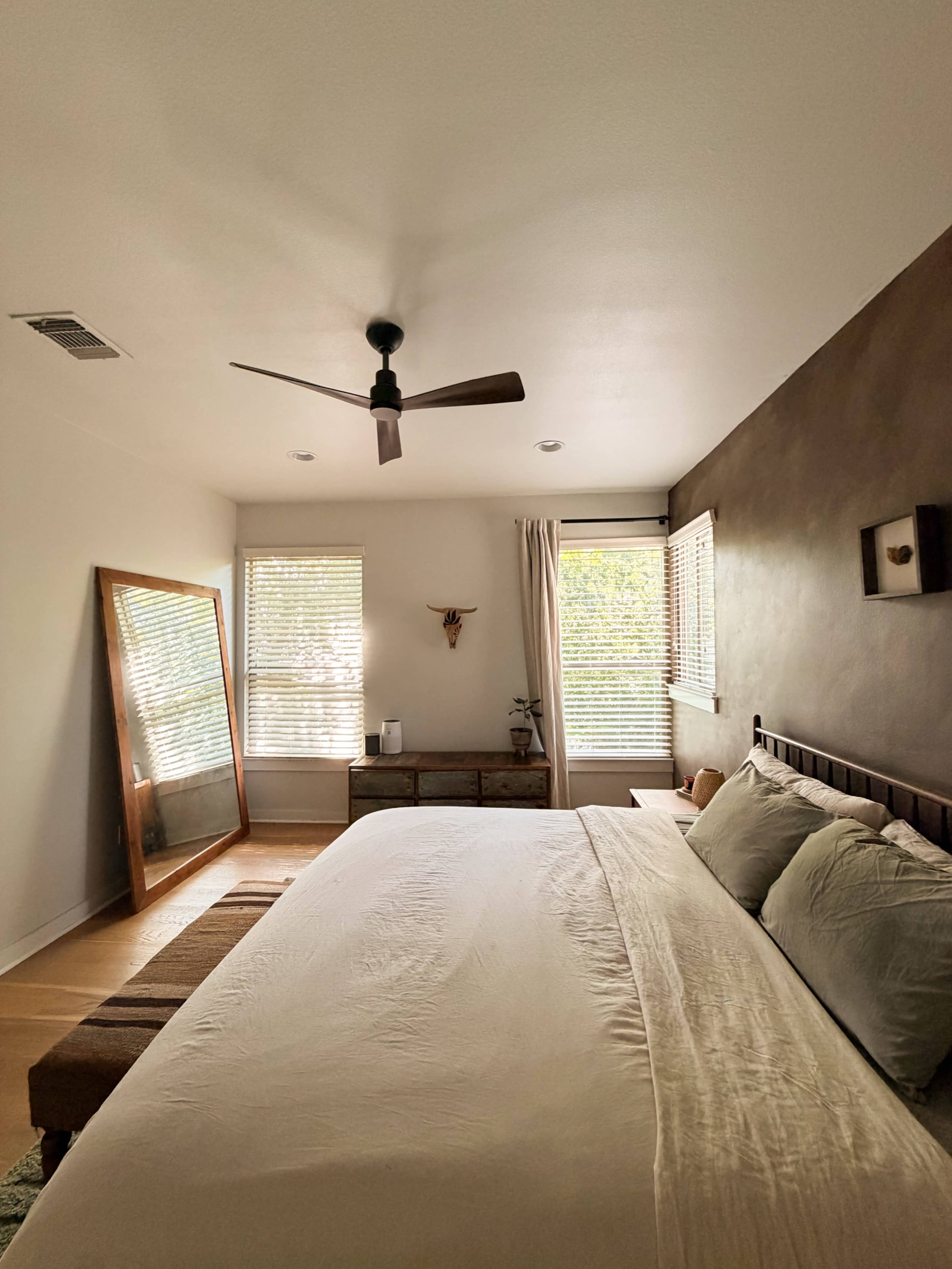 The image shows a modern bedroom with a large bed, a wooden bench at the foot, a mirror against the wall, and natural light coming through the window.