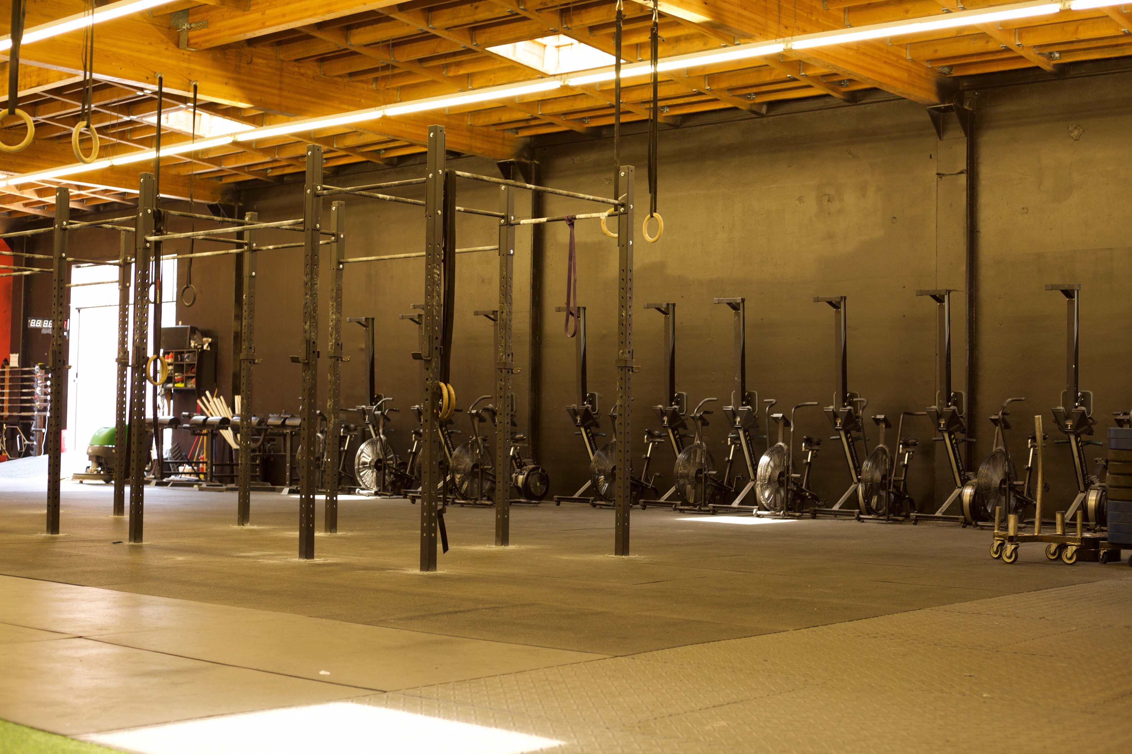 The image shows a spacious gym with multiple gymnastic rings and exercise equipment lined up against a dark wall under wooden beams.