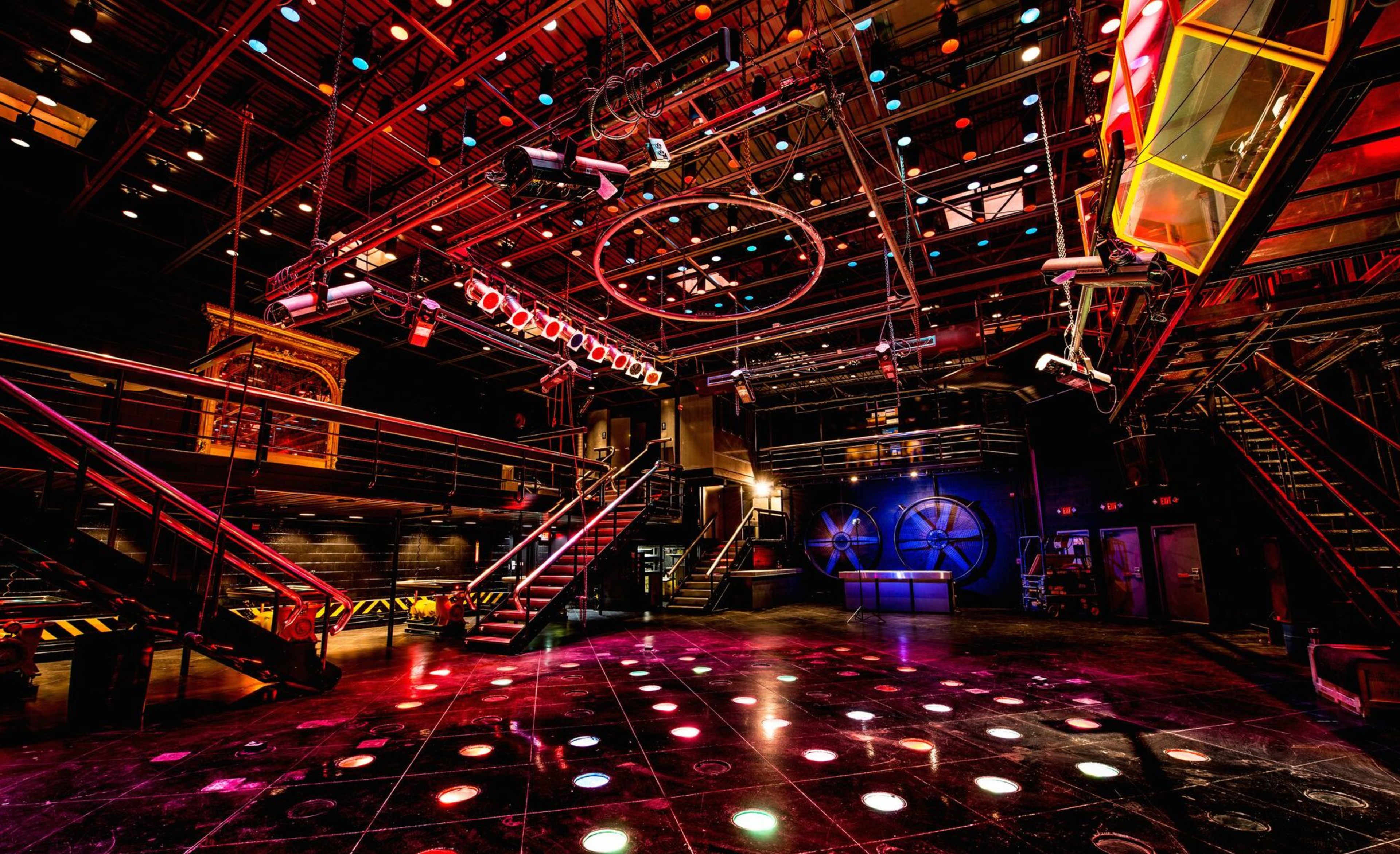 A spacious, illuminated theater or performance venue with a polished floor, multicolored lights, and multiple levels with staircases.
