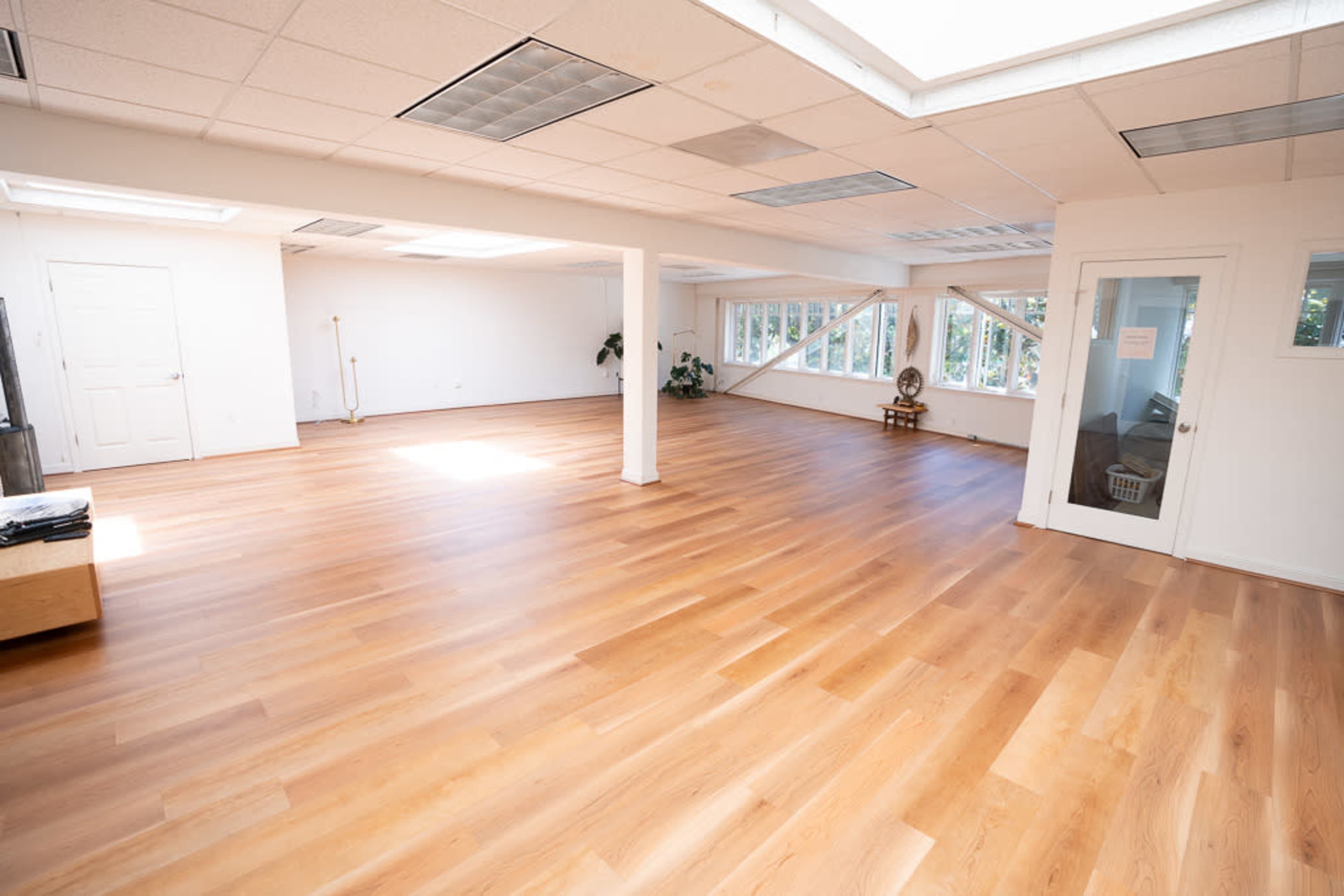 A spacious, brightly lit room with wooden floors and large windows, featuring minimal furnishings.