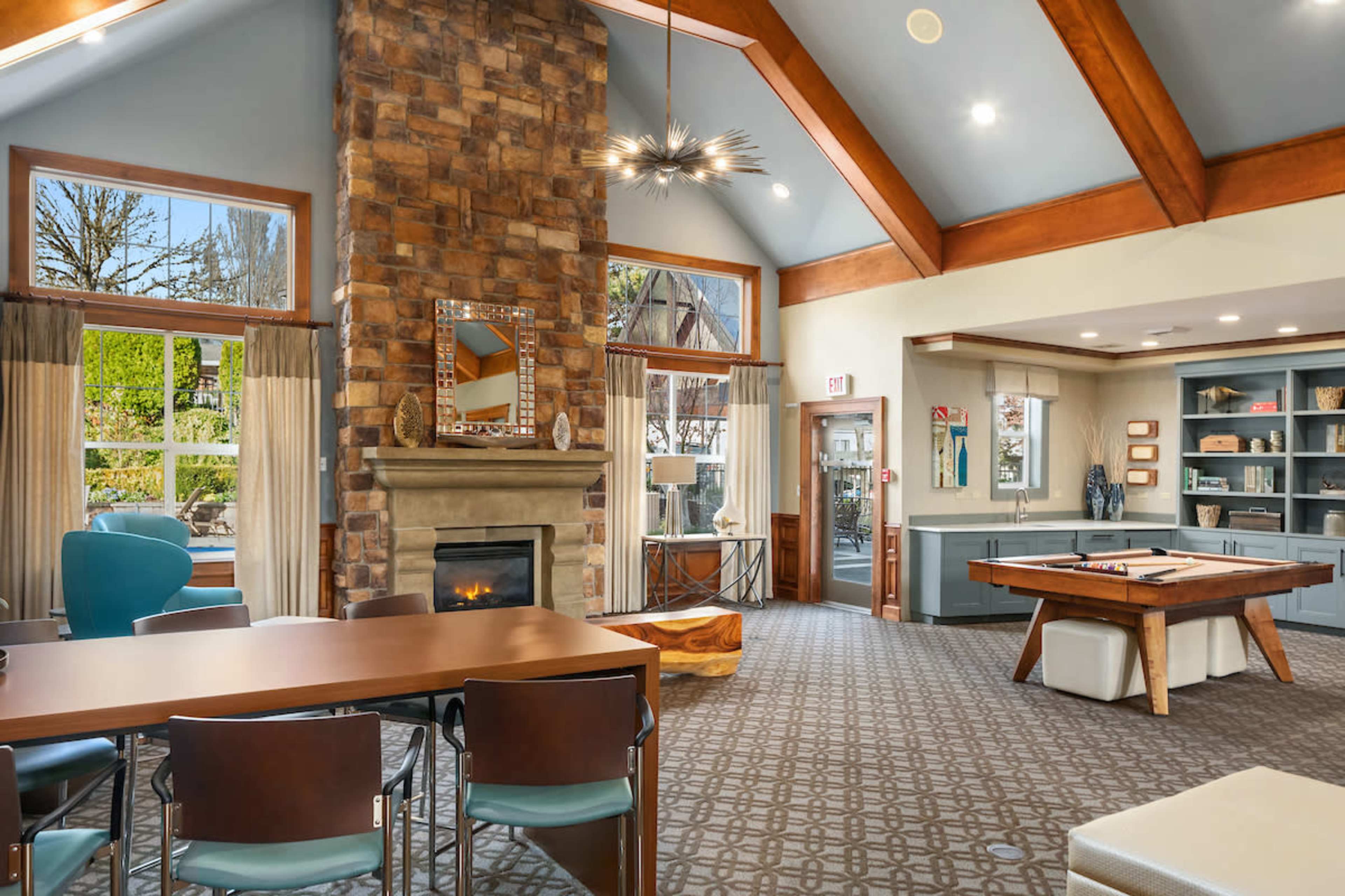 The image shows a modern community lounge area featuring a stone fireplace, large windows with views of greenery, a pool table, and a dining table with chairs.