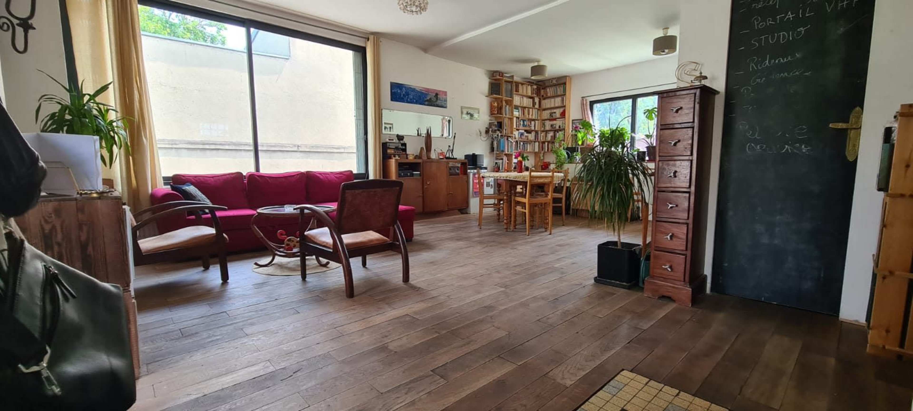 The image shows a cozy living room with wooden flooring, featuring a red sofa, a wooden dining table, and bookshelves filled with various items.