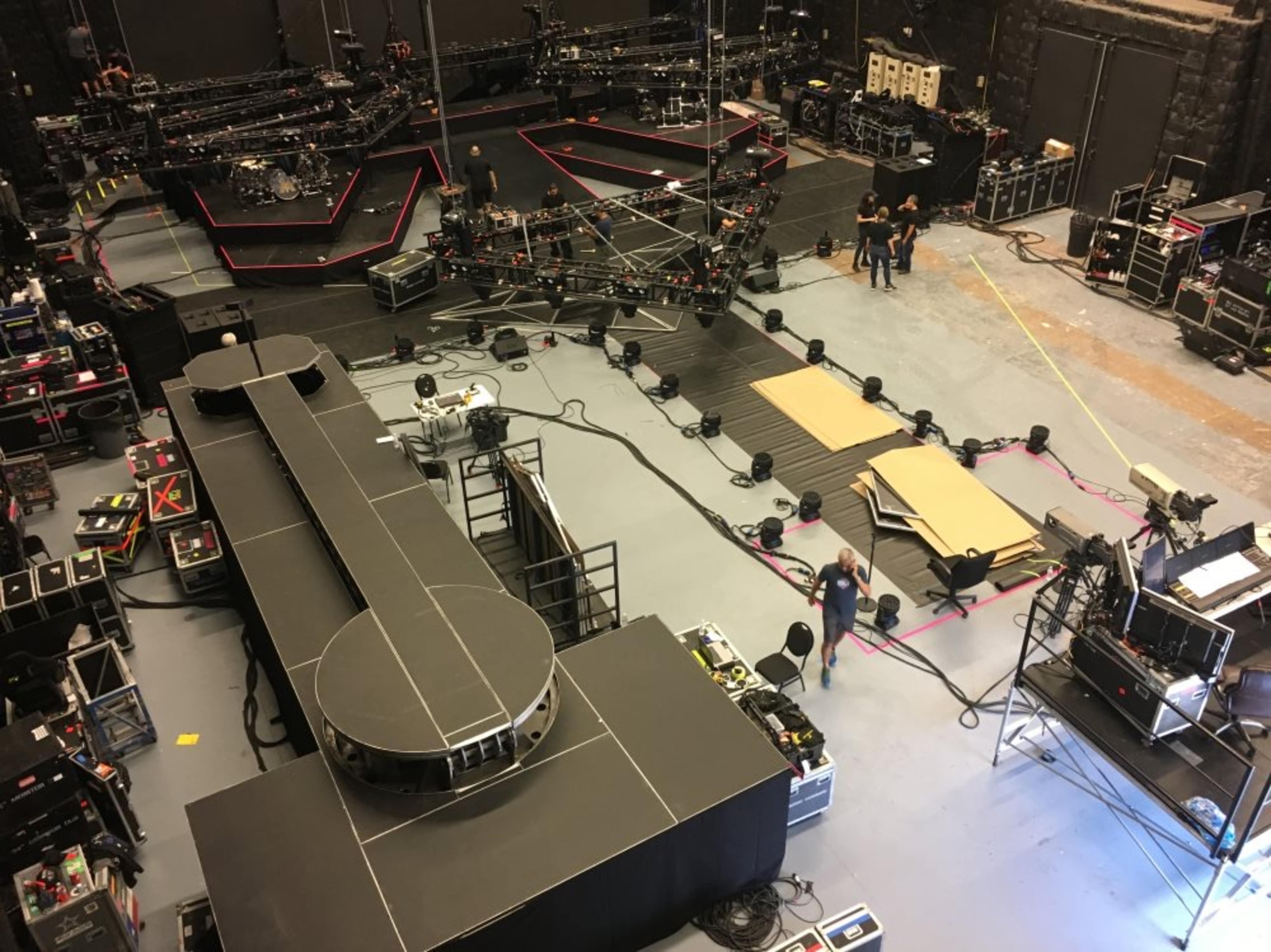 A bird's-eye view of a concert stage setup with various equipment, lighting rigs, and wooden platforms arranged in a rehearsal space.