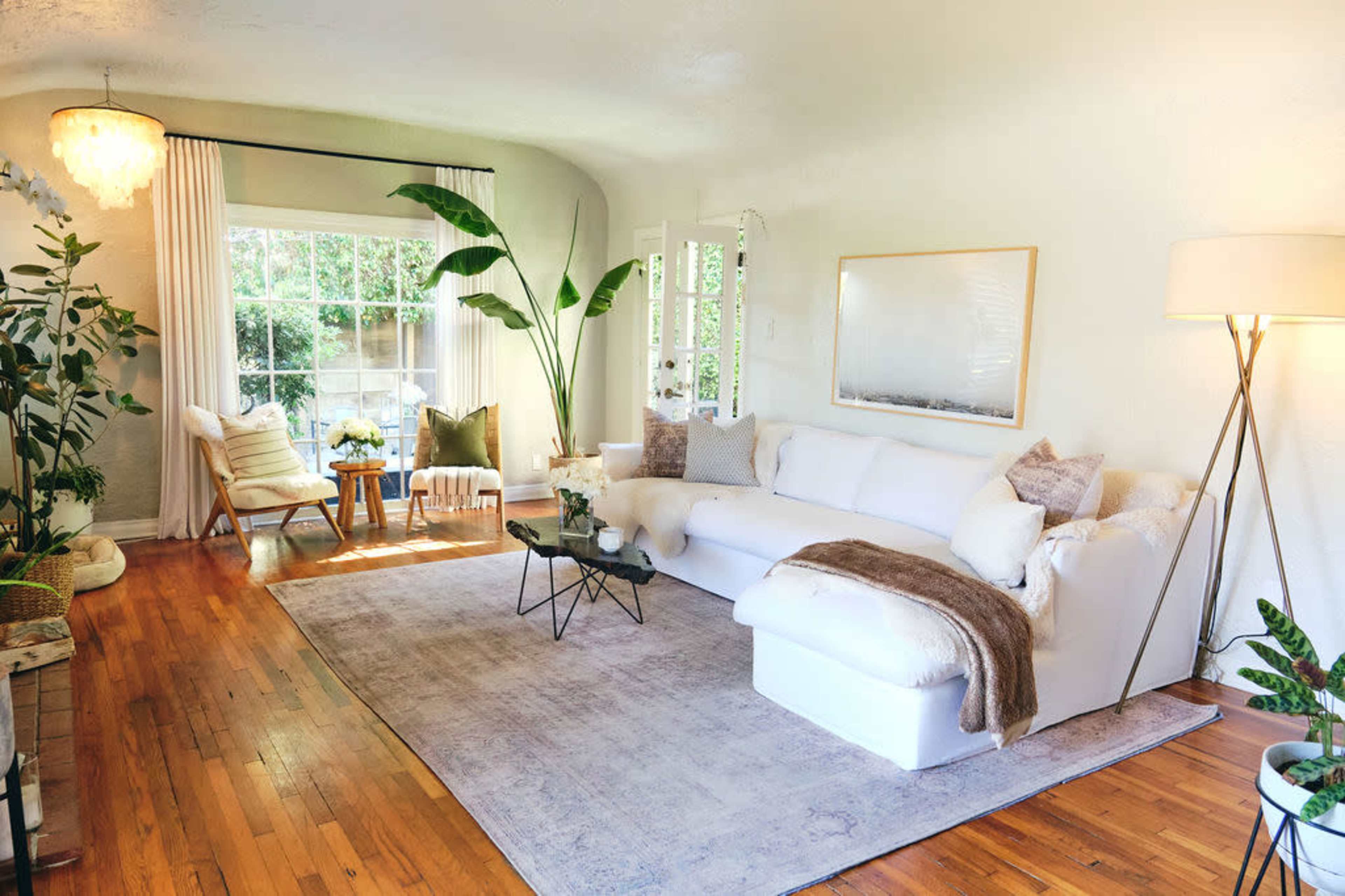 The living room features a white sectional sofa, a coffee table, and large windows allowing natural light, surrounded by greenery and decorative items.