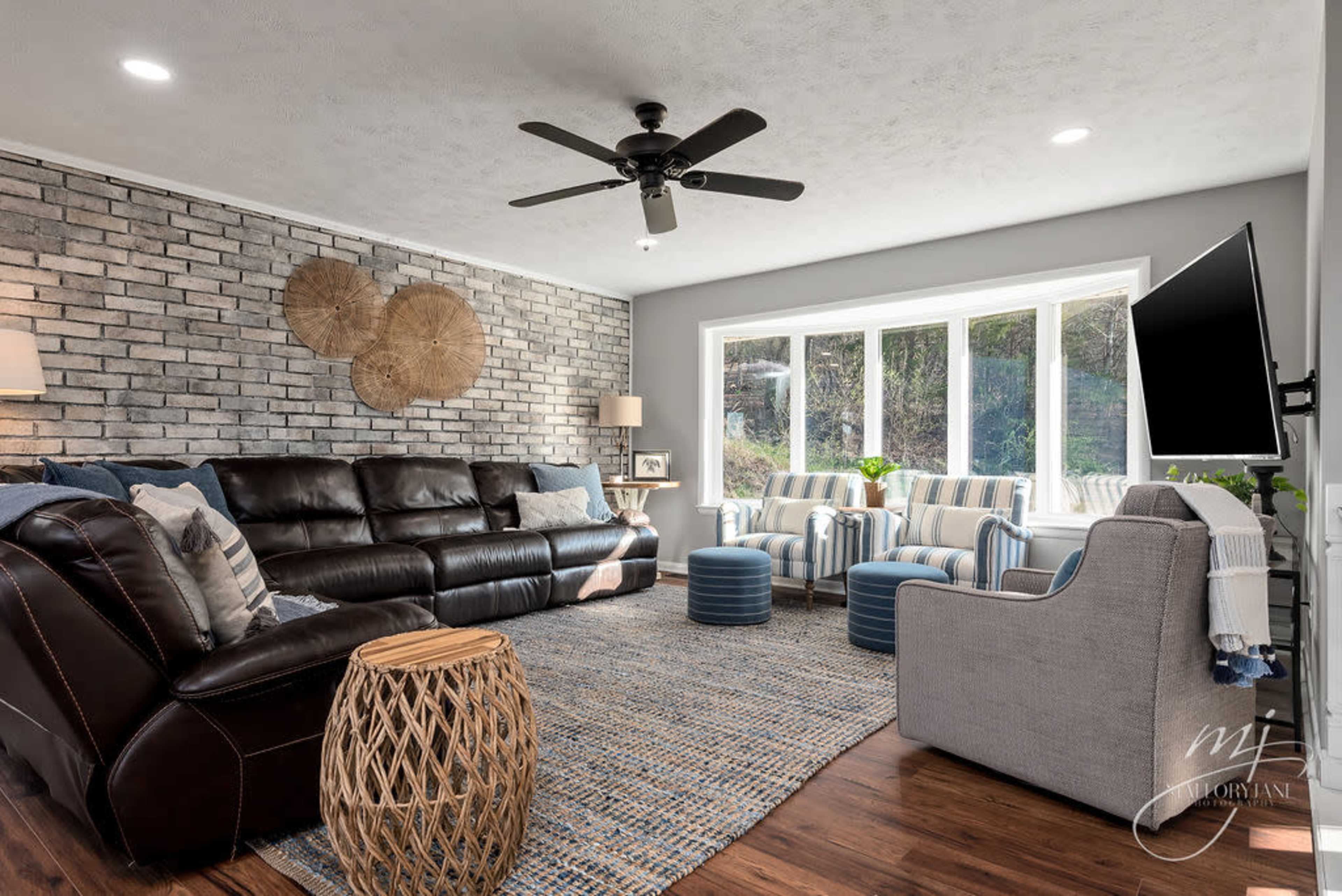 The living room features a combination of a brick wall, a dark leather sofa, two striped chairs, and a round coffee table, all illuminated by natural light from large windows.