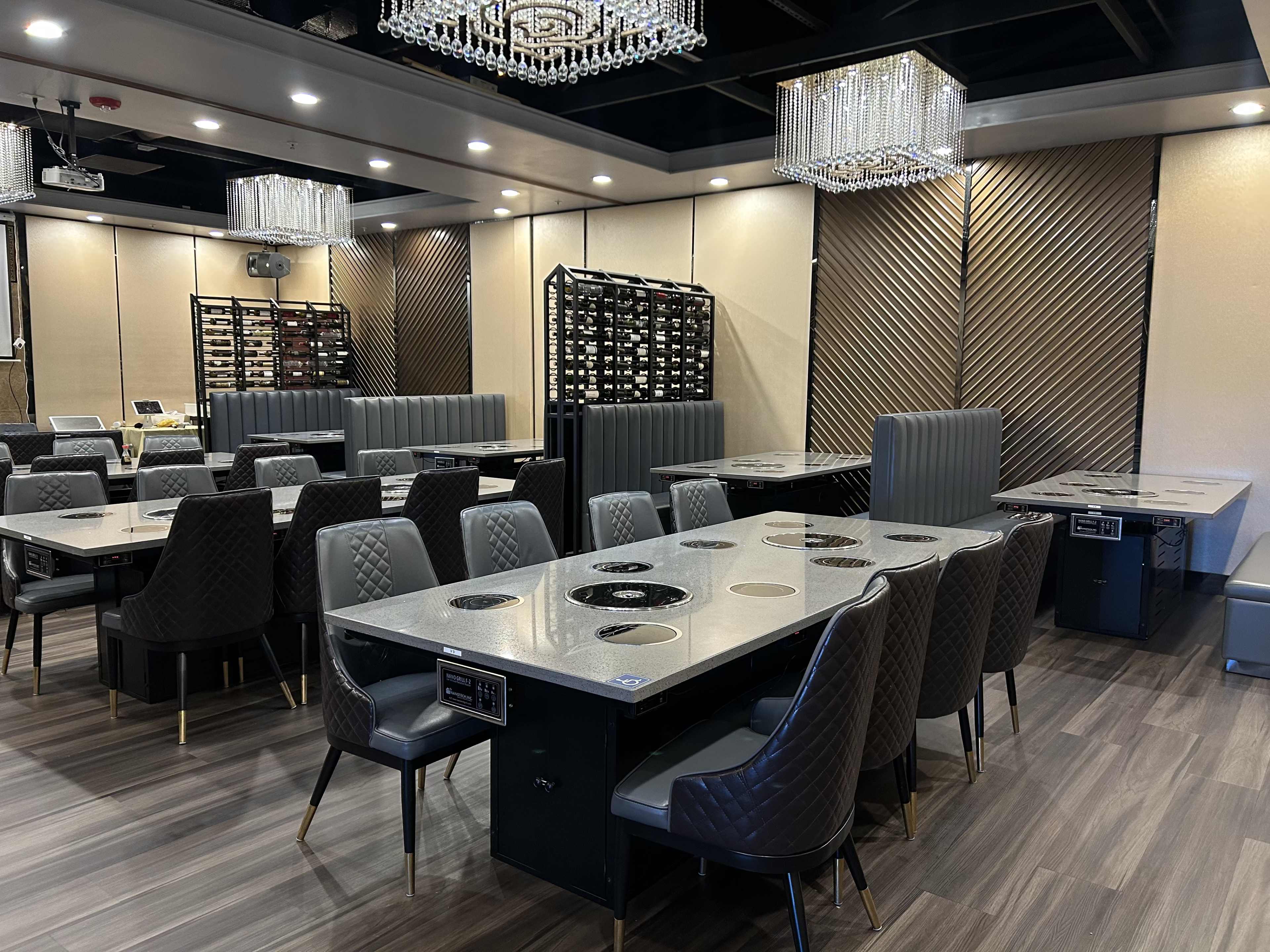A modern dining area with several gray tables equipped for hot pot cooking, surrounded by plush black chairs and adorned with decorative chandeliers.