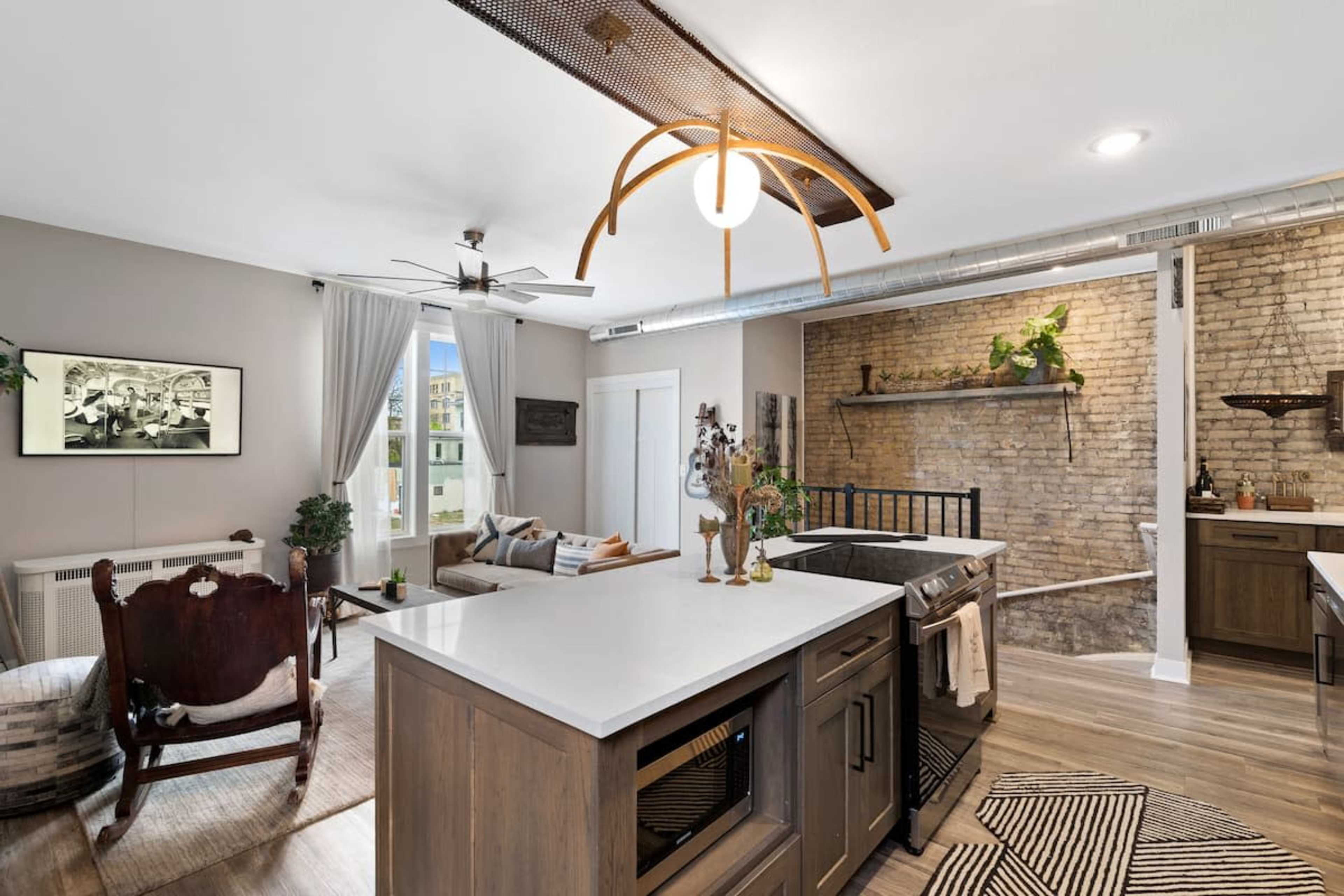 The image features a modern kitchen with contrasting wooden cabinetry, a central island, and a brick wall, adjacent to a cozy living area with a vintage chair and stylish decor.