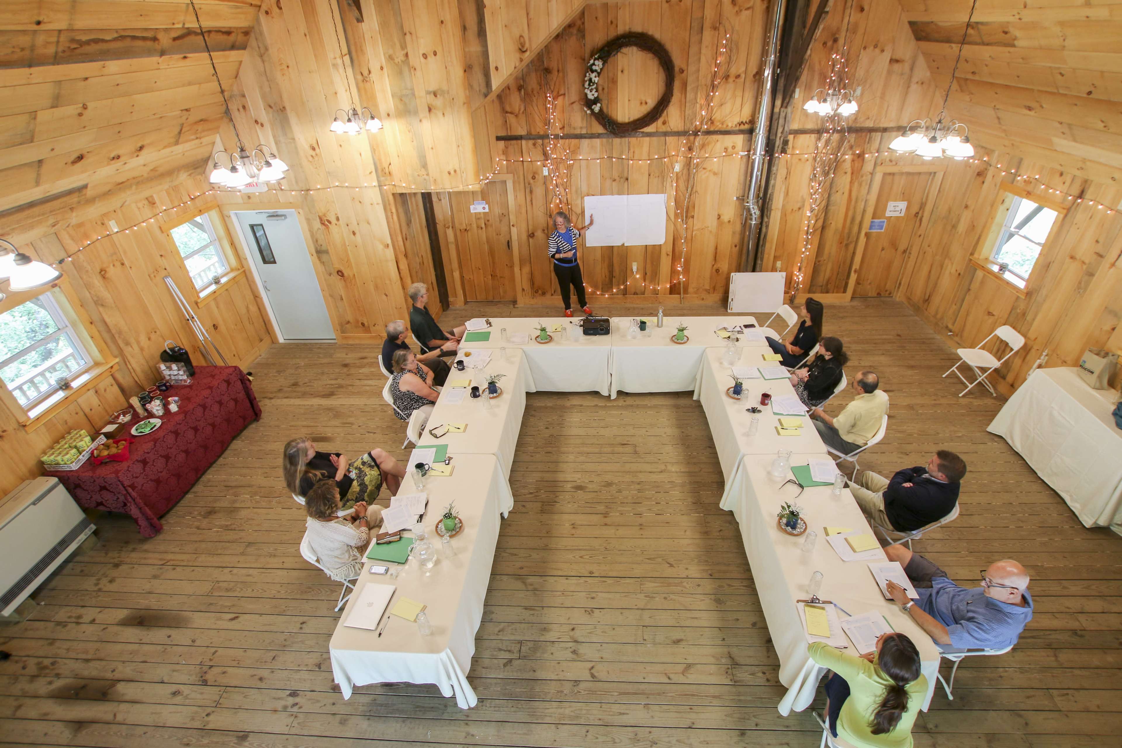 A group of people is seated around a long table in a wooden meeting room while a presenter stands at the front by a whiteboard.
