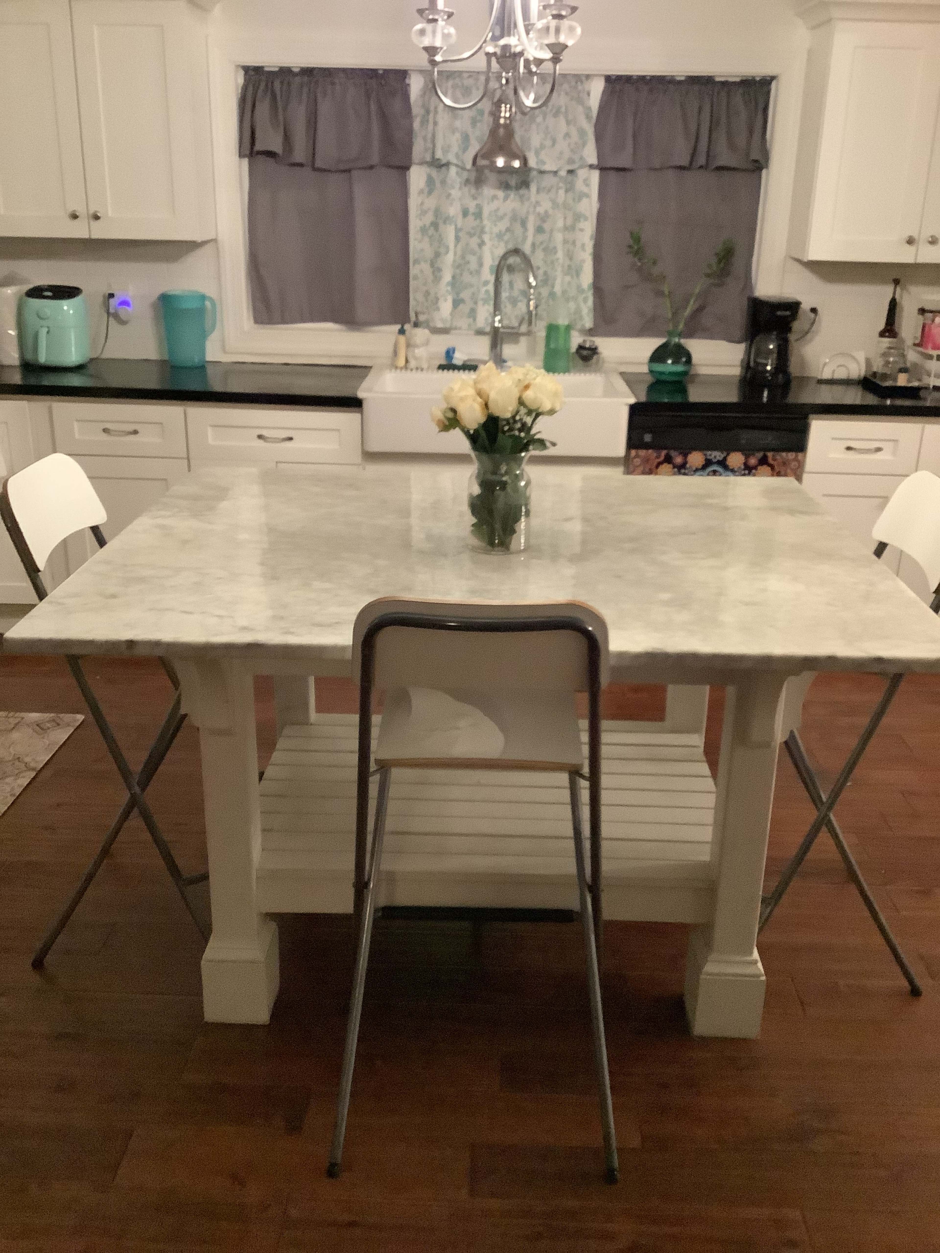 A marble-top table with a vase of flowers is surrounded by four chairs in a kitchen with white cabinetry and dark wood flooring.