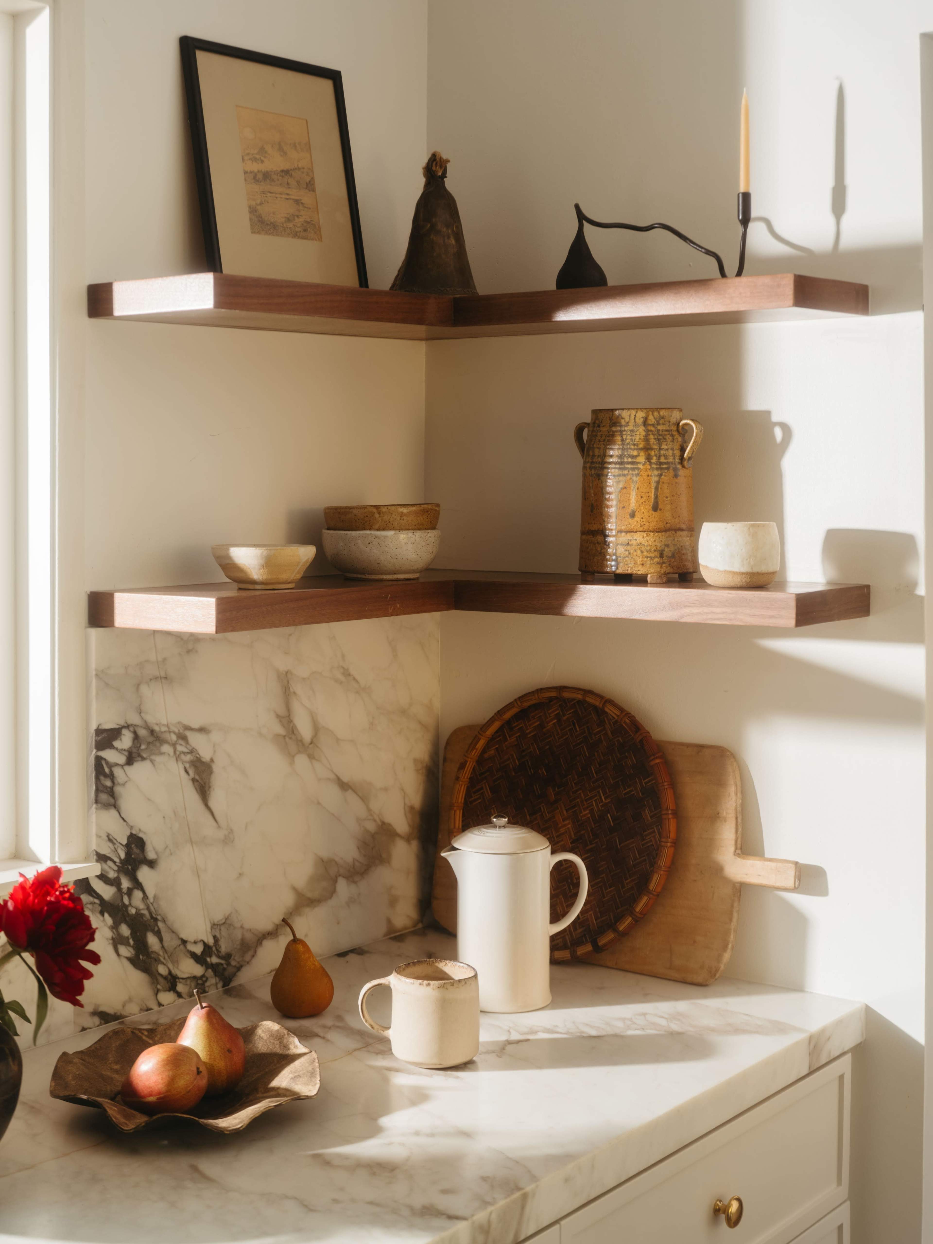 The image shows a corner kitchen shelf with wooden shelves displaying various ceramic bowls and a pitcher, alongside a marble countertop featuring a woven basket and a cutting board.