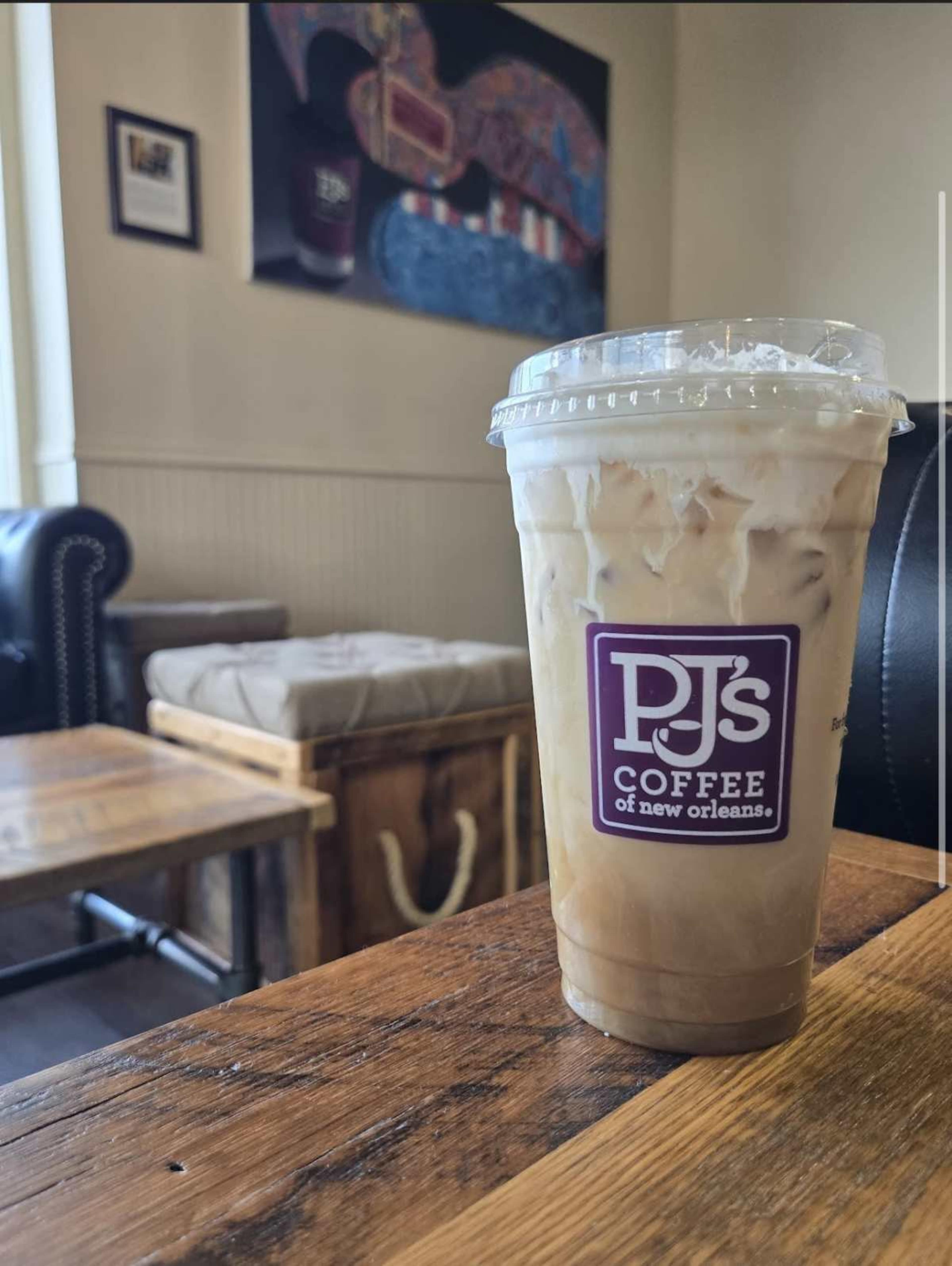 A clear cup of iced coffee from PJ's Coffee sits on a wooden table in a café with a cozy interior.