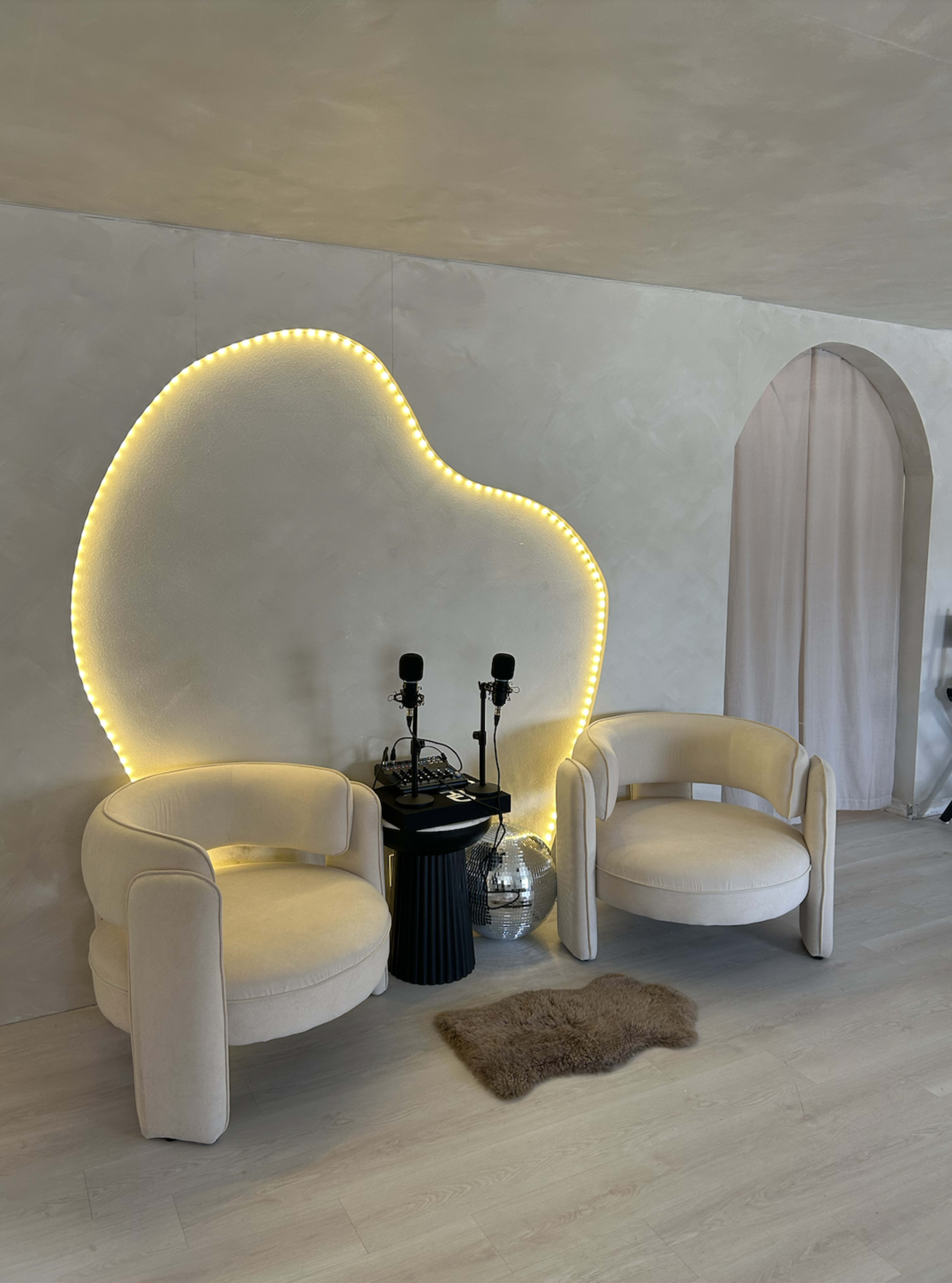 The image shows two cream-colored chairs positioned facing a wall with a unique, illuminated design and two microphones on a black pedestal beside them.