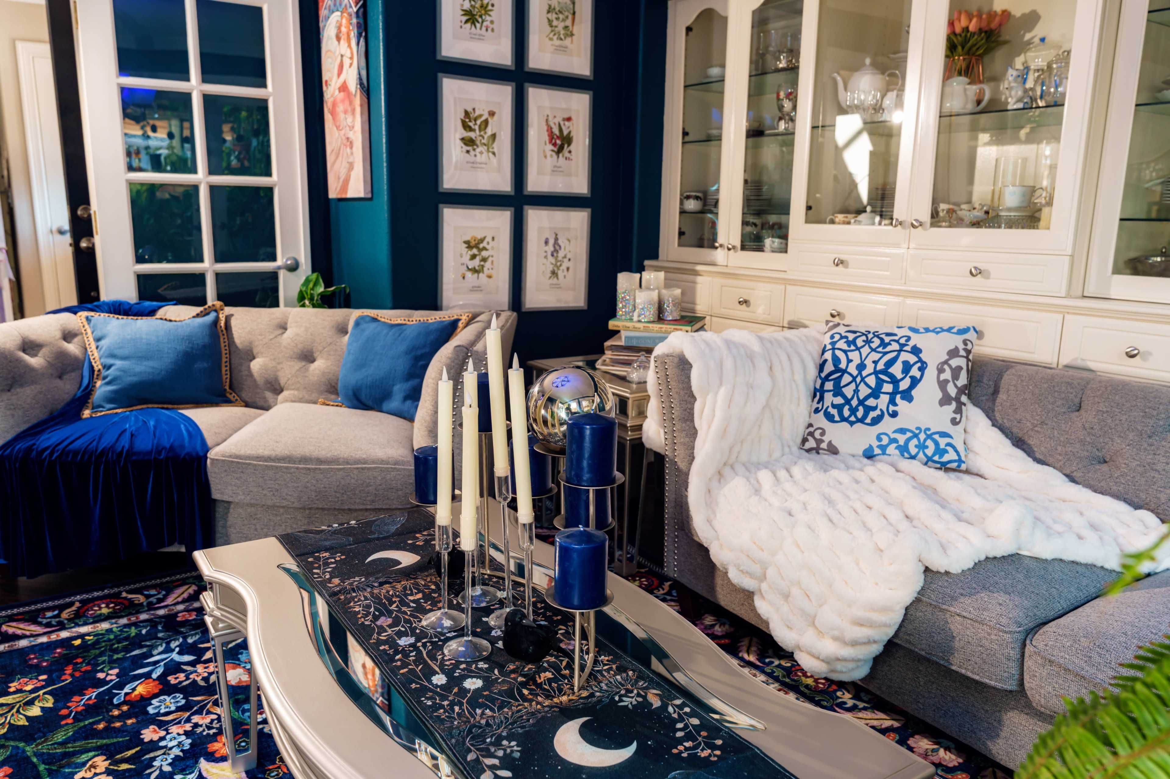 The image shows a cozy living room with two gray sofas adorned with blue and white pillows, a decorative table with candles, and a patterned rug.