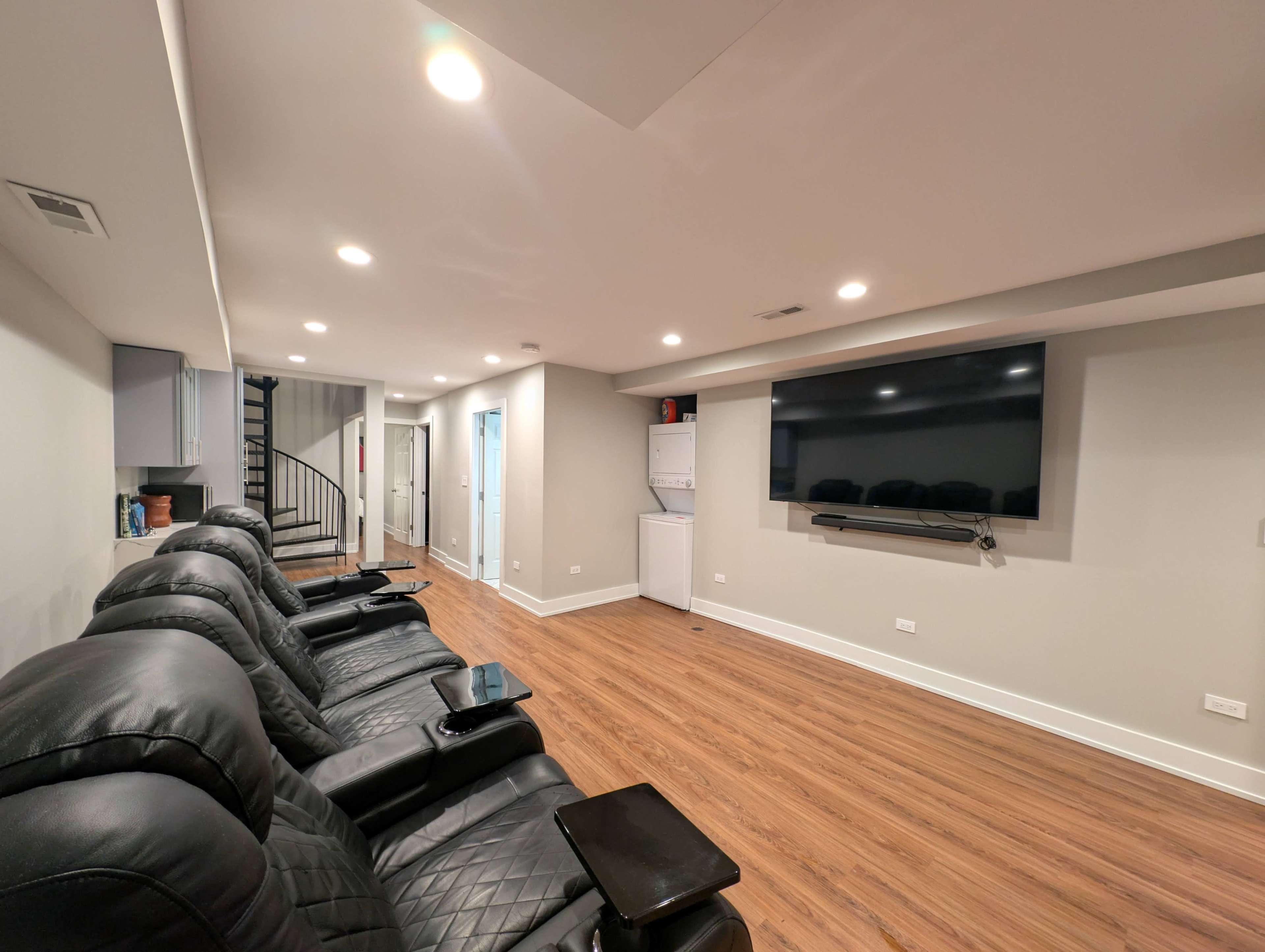 The image shows a spacious room with wooden flooring, featuring black reclining chairs facing a large wall-mounted television and a spiral staircase in the corner.