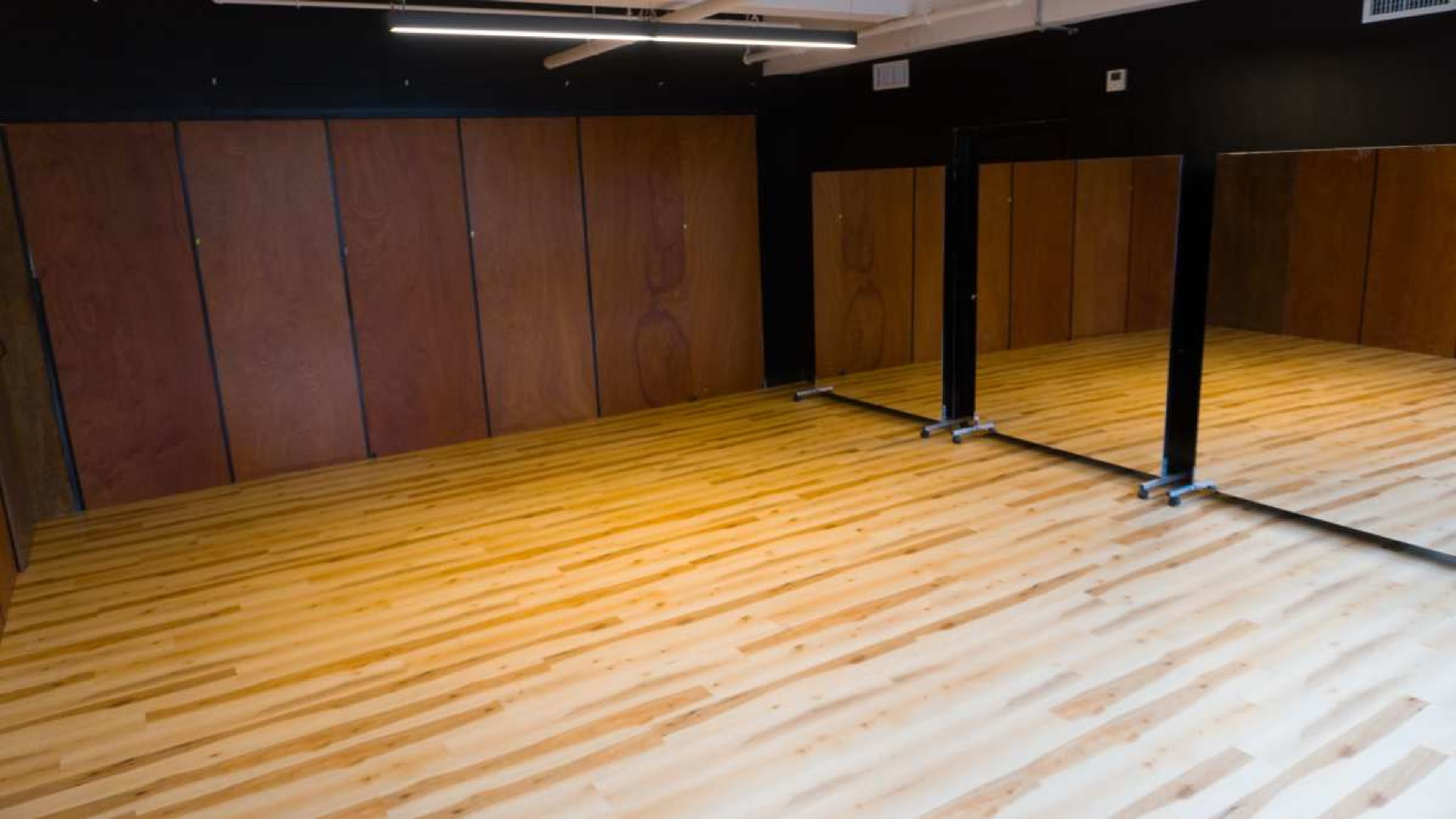 The image shows a spacious, empty room with wooden flooring and large mirrors on one side, complemented by dark wood paneling on the walls.