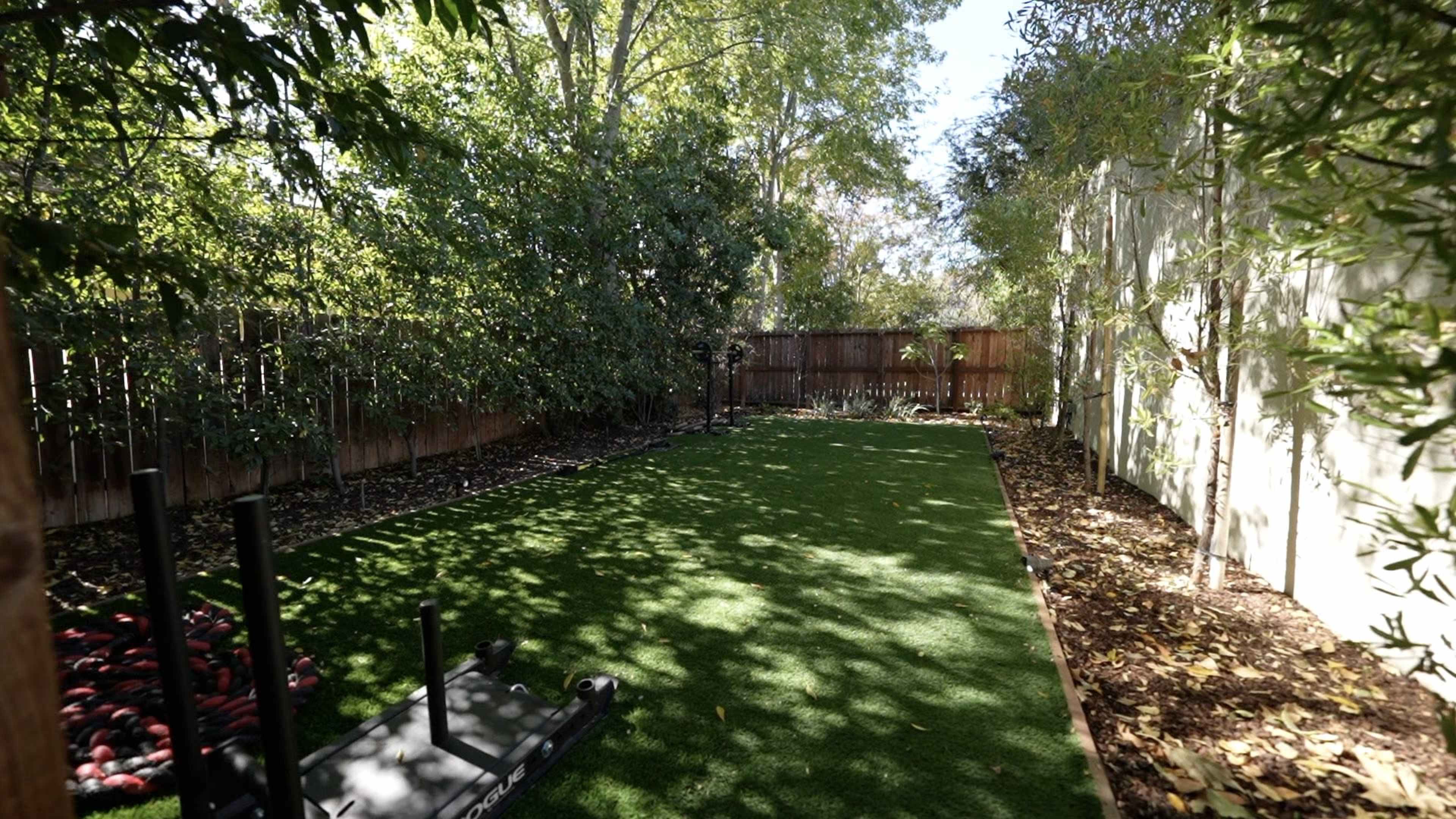 A rectangular backyard area with artificial turf, surrounded by trees and a wooden fence.