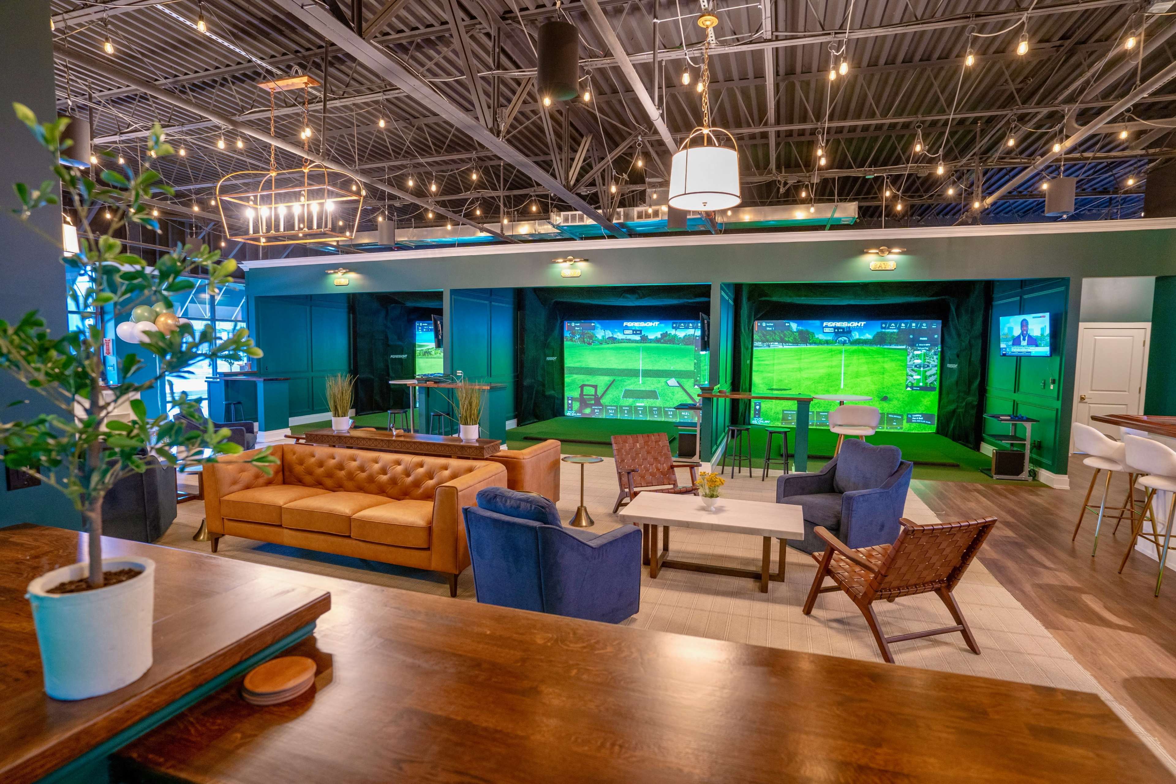 The image shows a spacious indoor lounge area featuring multiple golf simulators, comfortable seating, and modern decor.