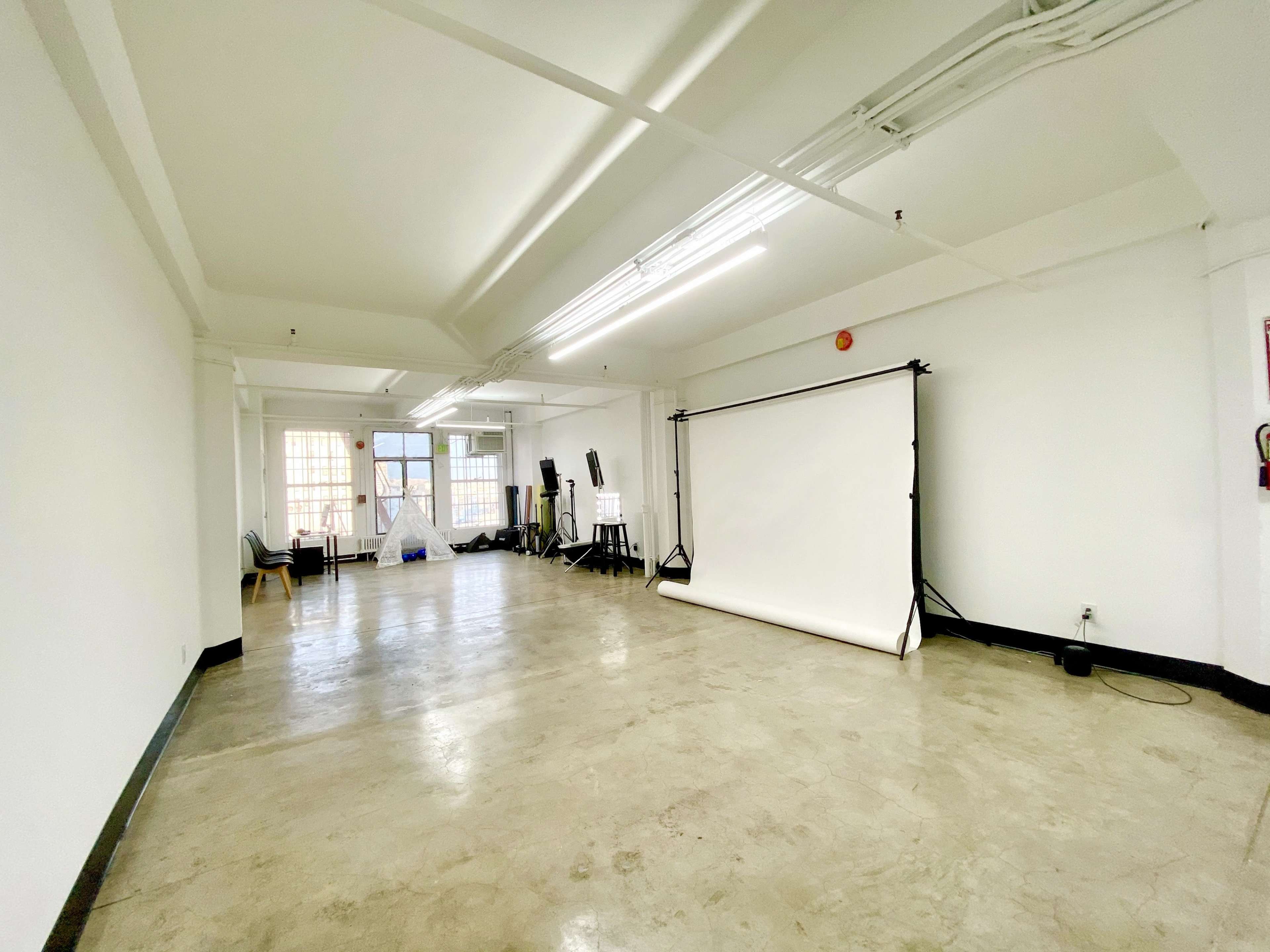 A spacious empty studio with concrete floors, white walls, and a backdrop setup at one end.