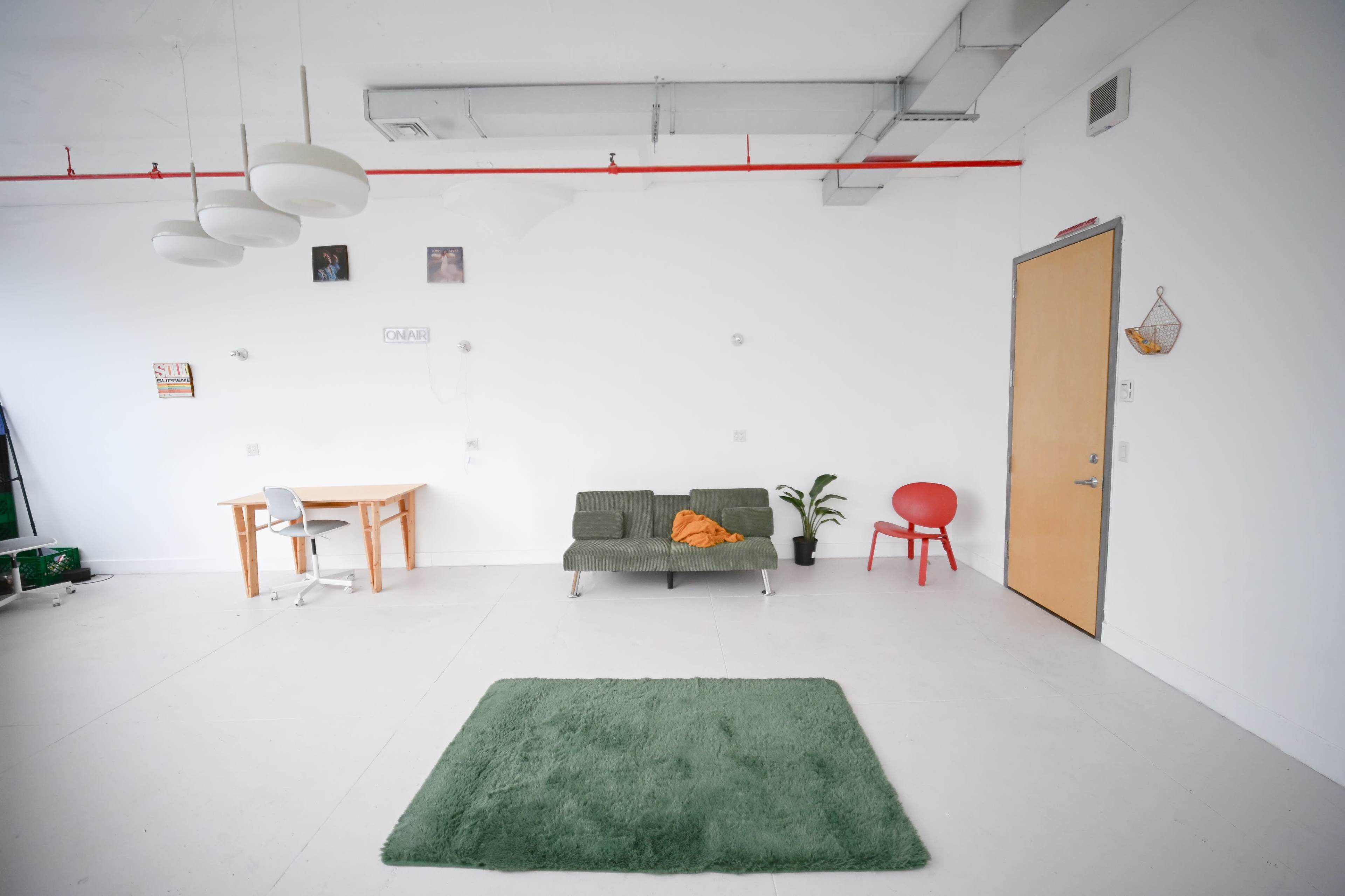 The image shows a minimalist room with a green rug, a gray sofa, a red chair, a wooden table, and simple decoration on the white walls.