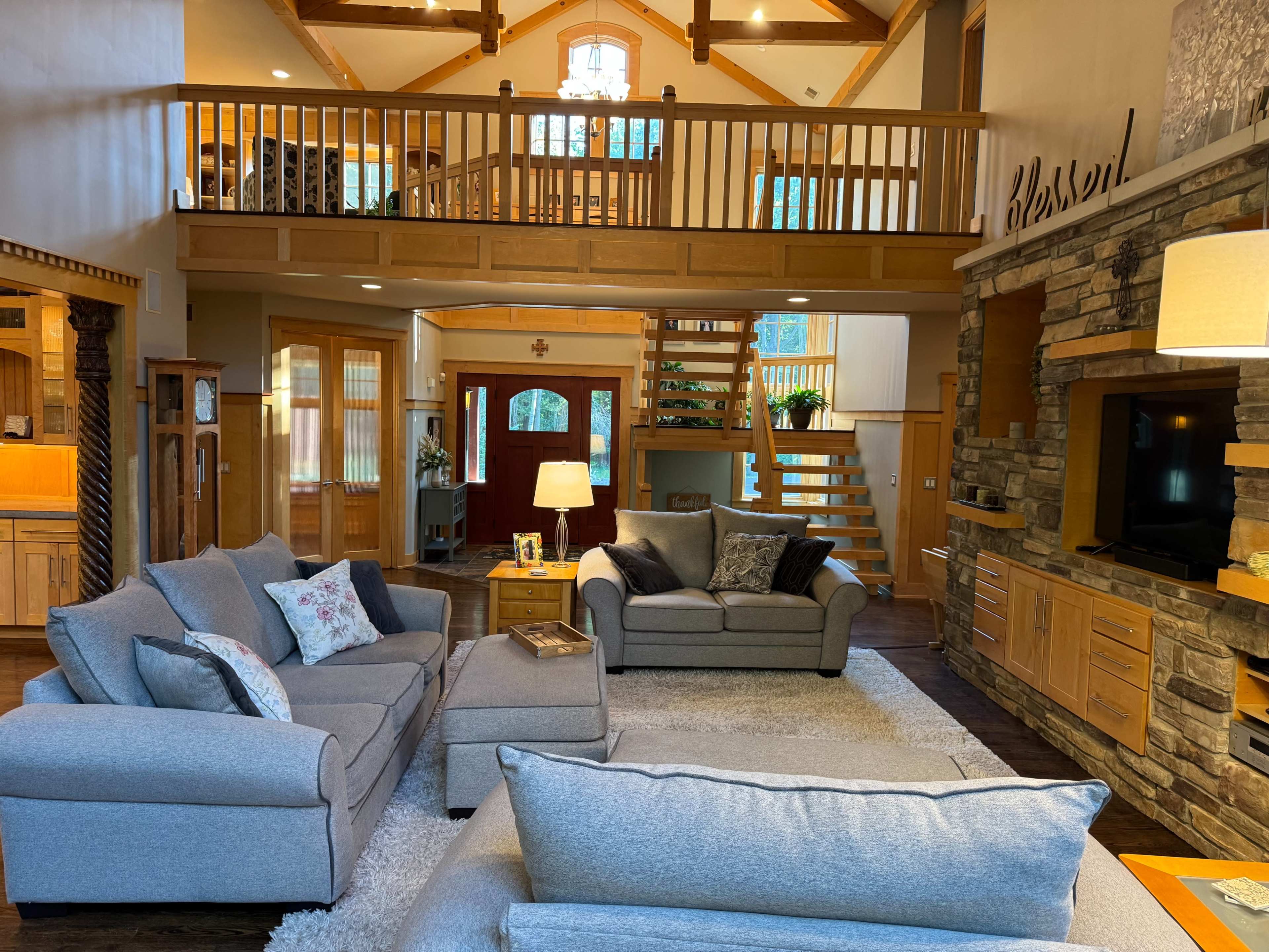 A spacious living room features comfortable gray sofas, a wooden staircase, and large windows allowing natural light to fill the space.