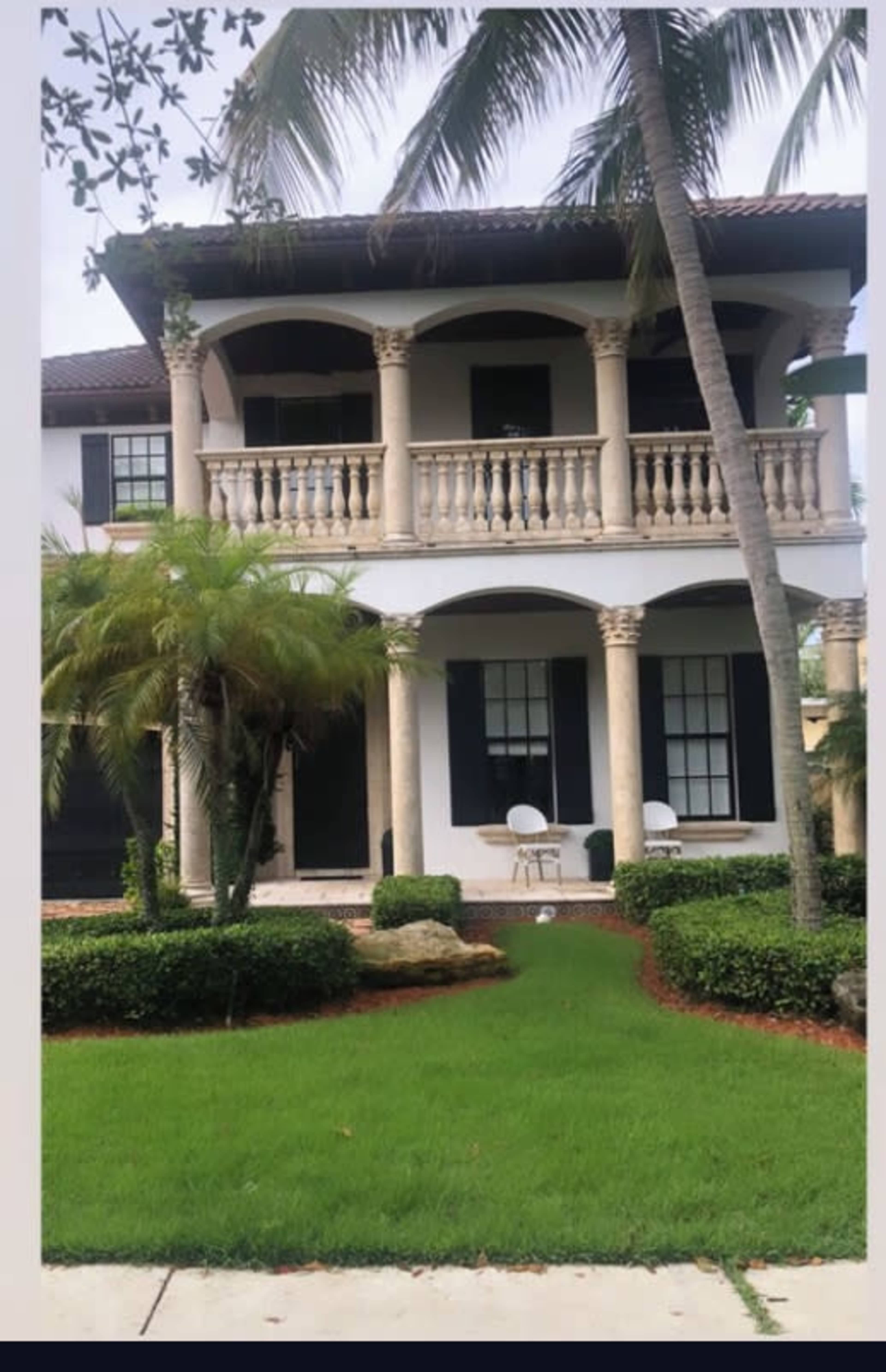 The image shows a two-story house featuring a balcony with columns, surrounded by a manicured lawn and palm trees.