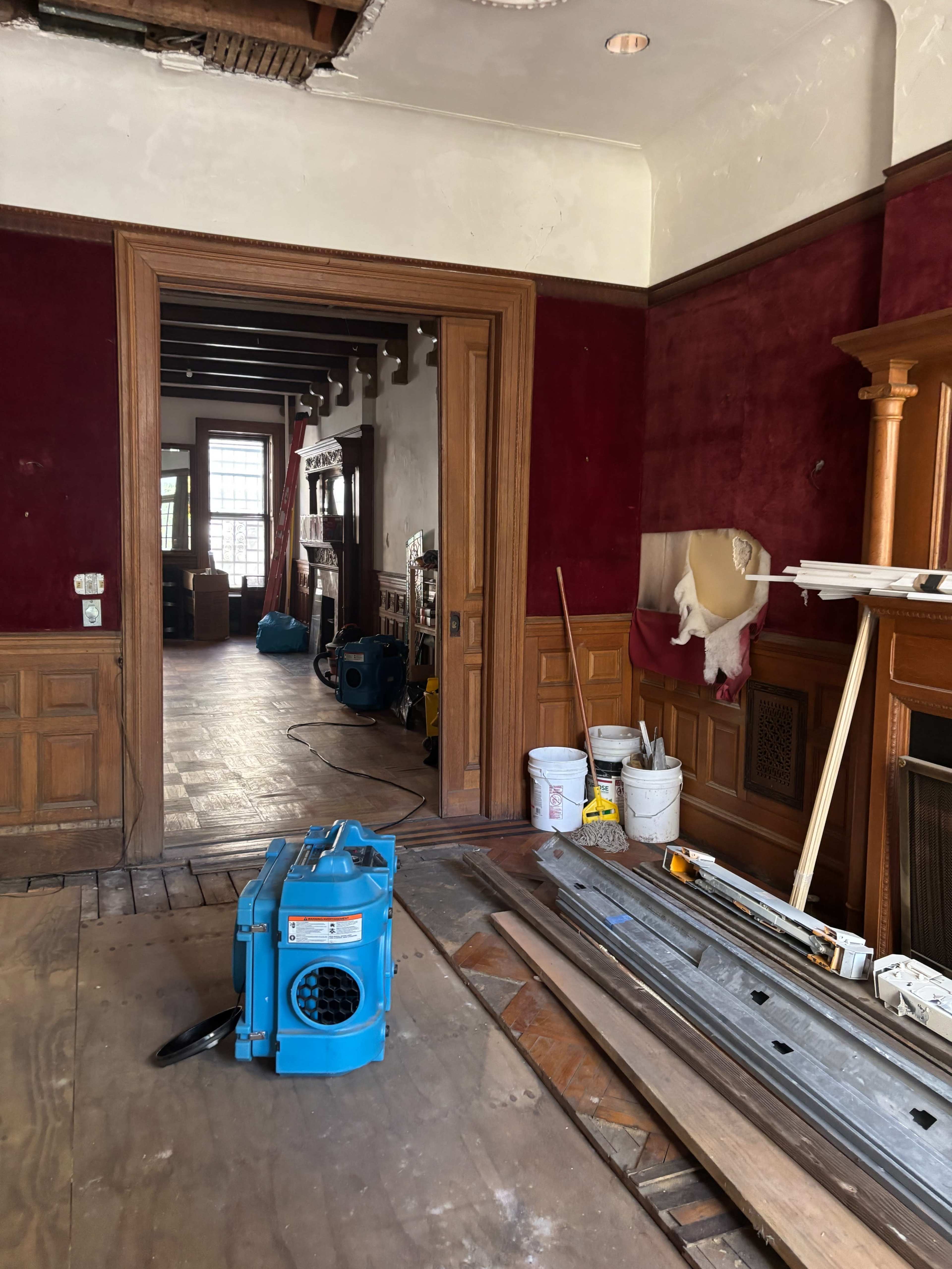 A room with red walls features a wooden floor, construction materials, and a blue air blower near an open doorway leading to another area.