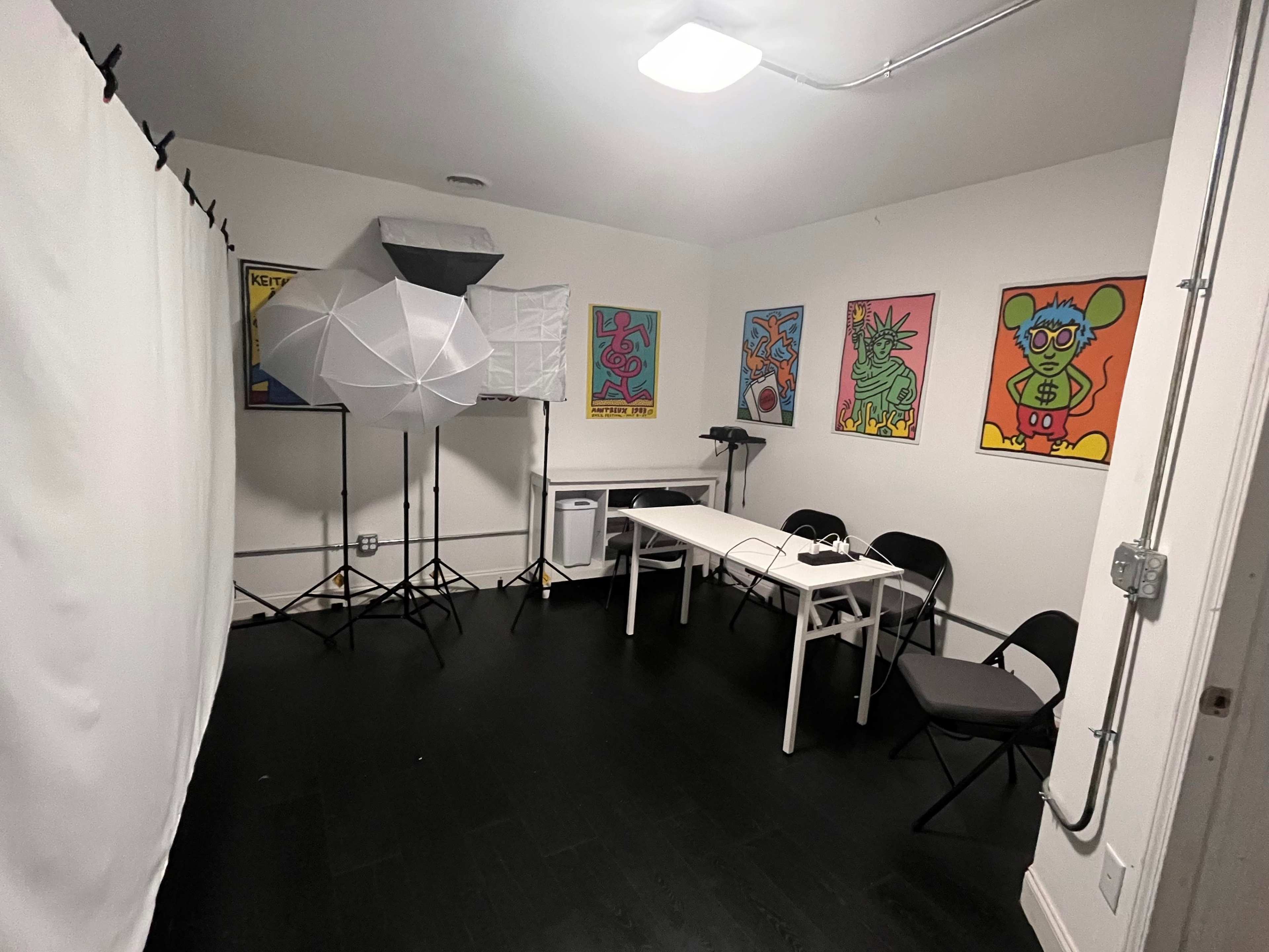 The image shows a small room with a white backdrop, a softbox lighting setup, a table, chairs, and colorful artwork on the walls.