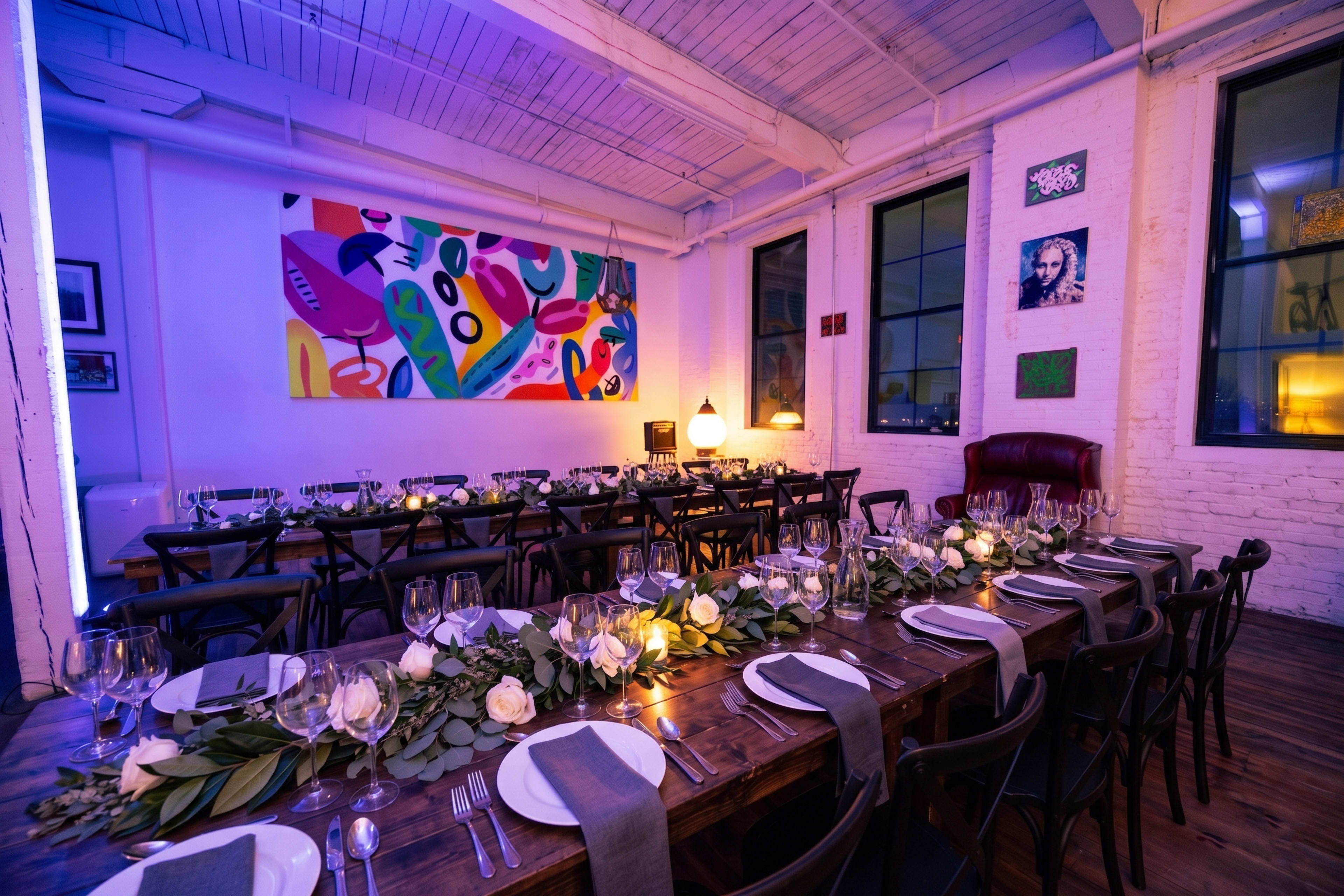 The image shows a spacious dining area set with long wooden tables, adorned with plates and glassware, surrounded by colorful artwork and softly lit by ambient lighting.