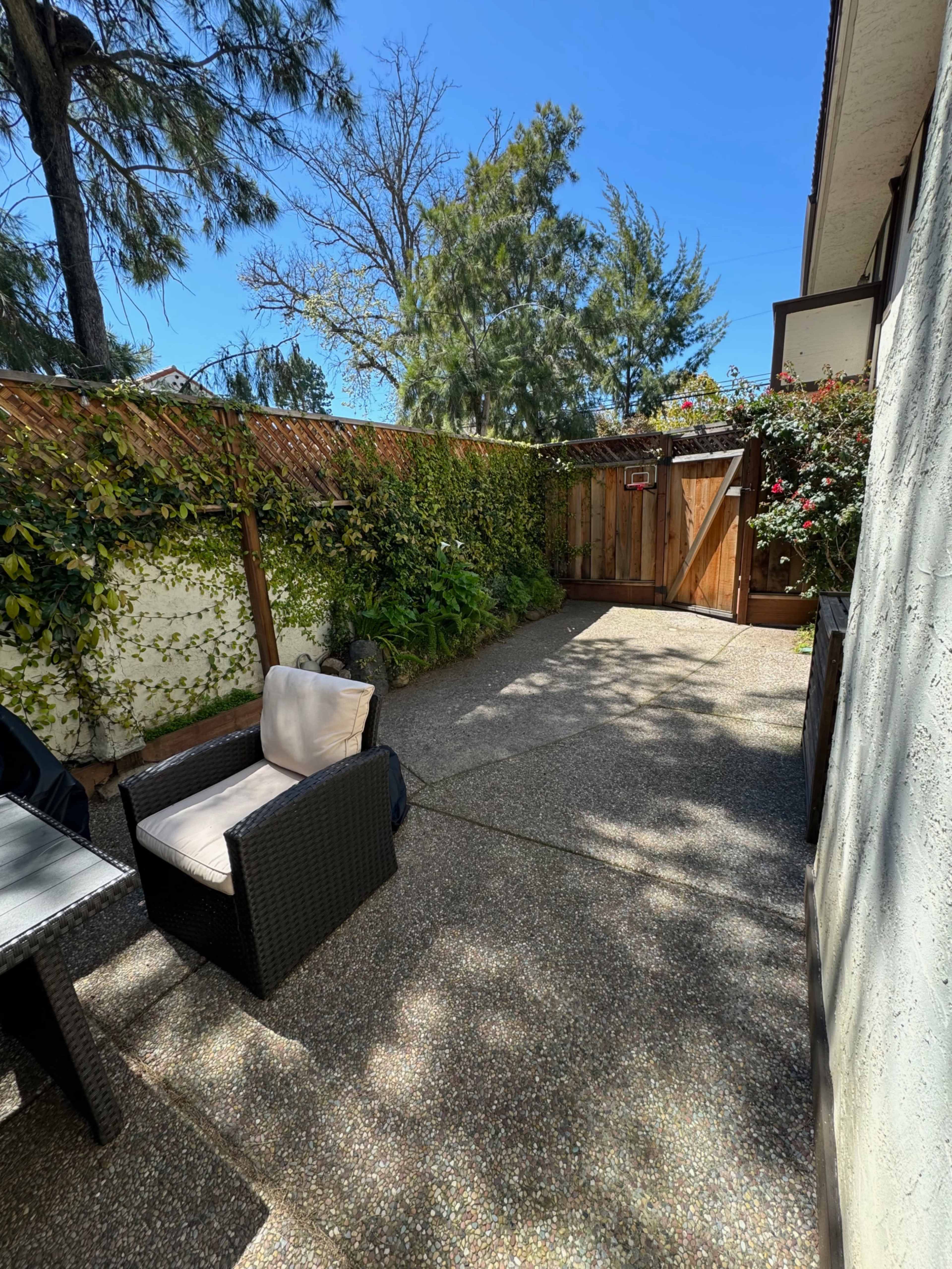 A small outdoor patio area with two chairs, a table, and a wooden gate surrounded by greenery and flowering plants.