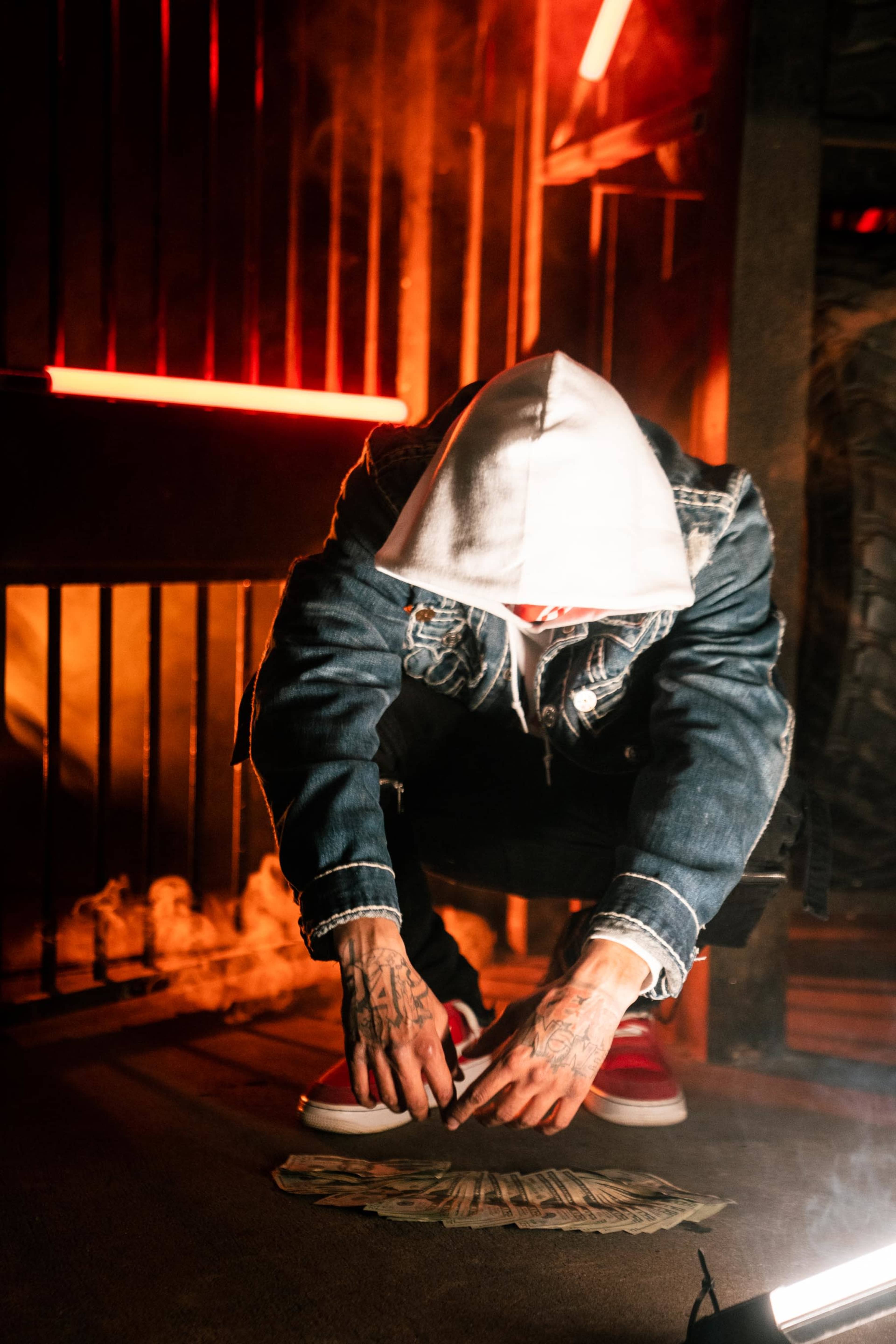 A person in a hoodie squats low to the ground, arranging stacks of cash in a dimly lit room with neon lighting and smoke.