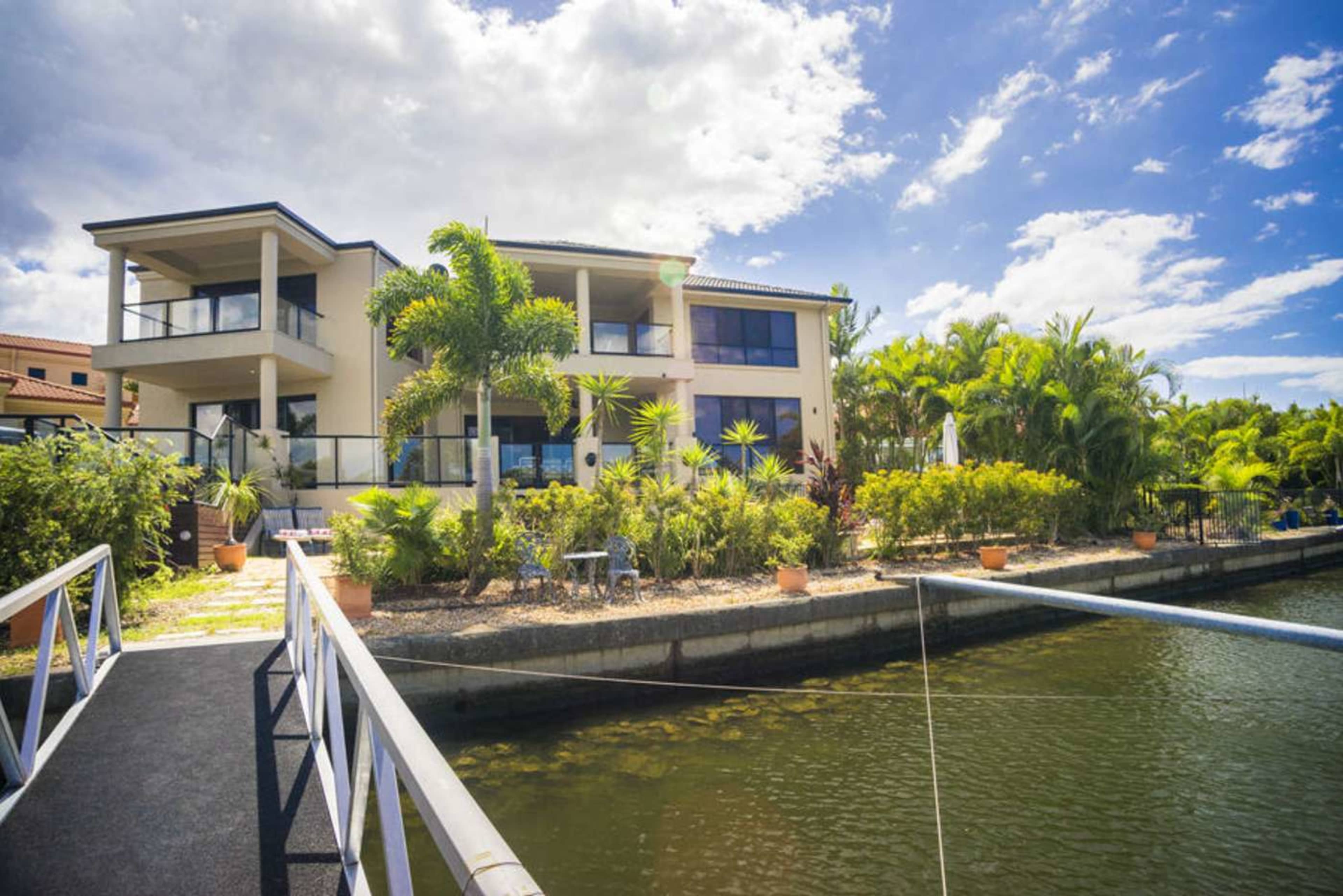 A modern two-story house with a balcony overlooks a landscaped garden and a canal.