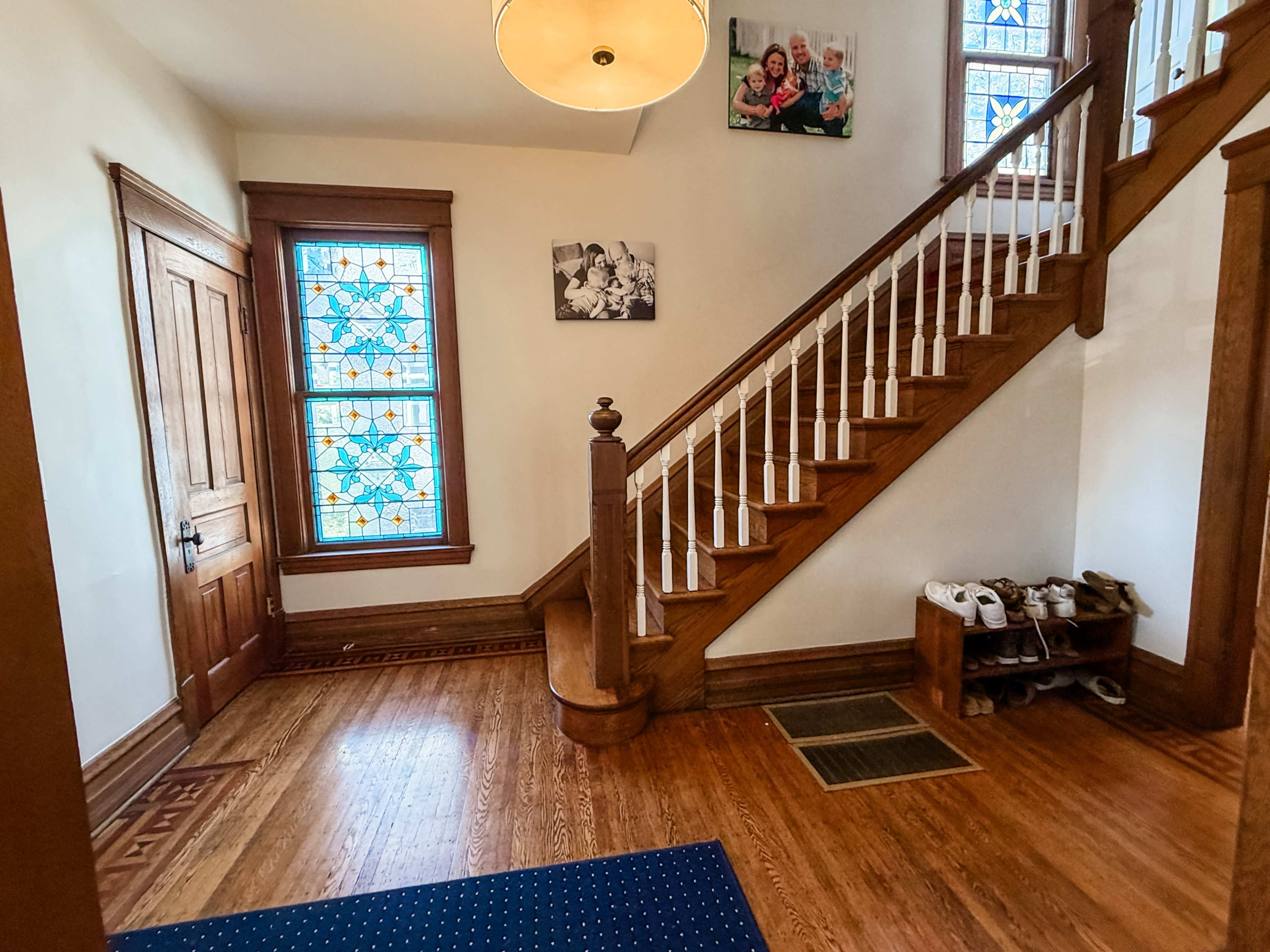 The image shows a foyer with a wooden staircase, stained glass windows, and a shoe rack near the entrance.