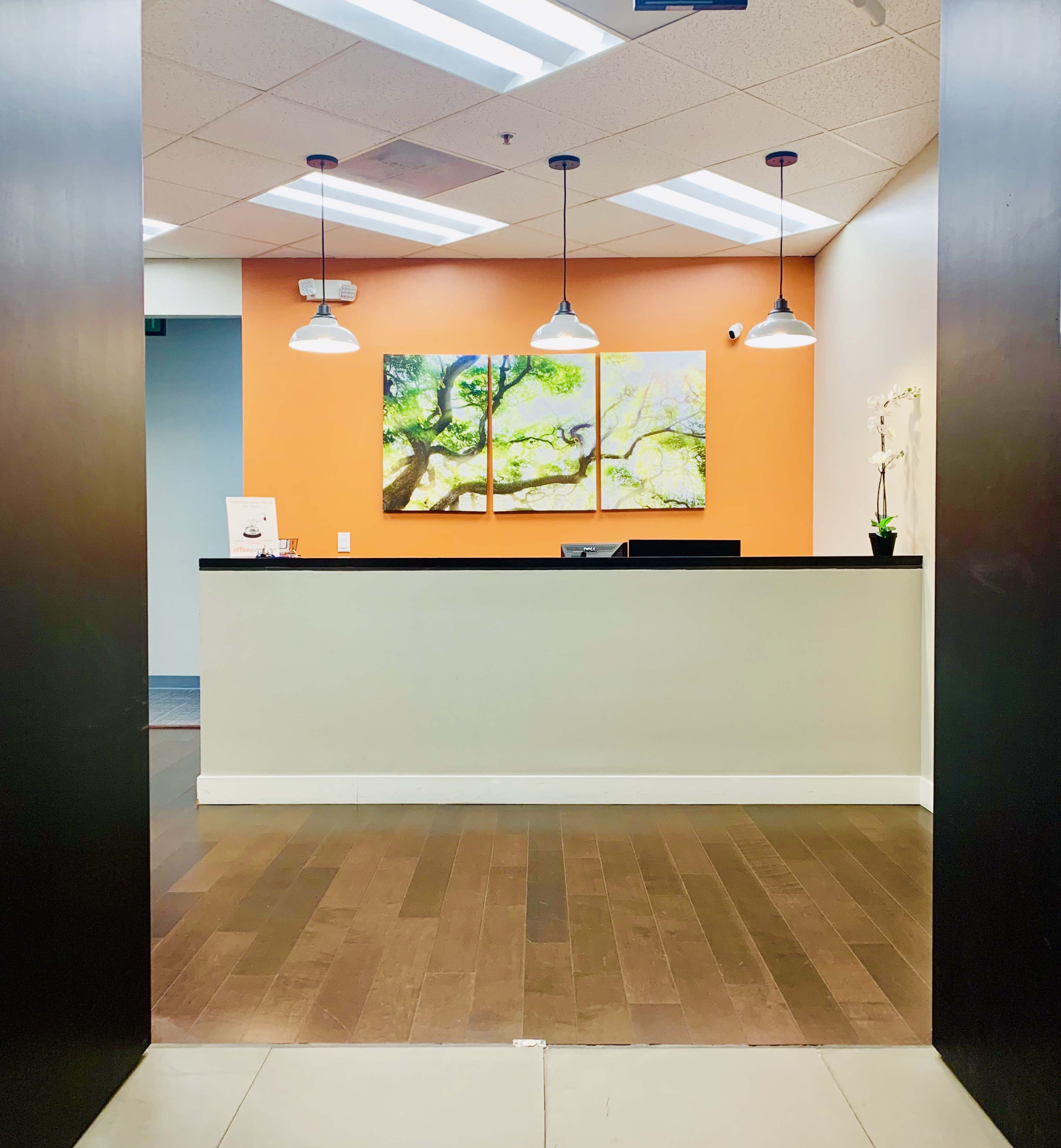 A reception area with a wooden floor, an orange wall, and three pendant lights above a counter topped with a framed artwork.