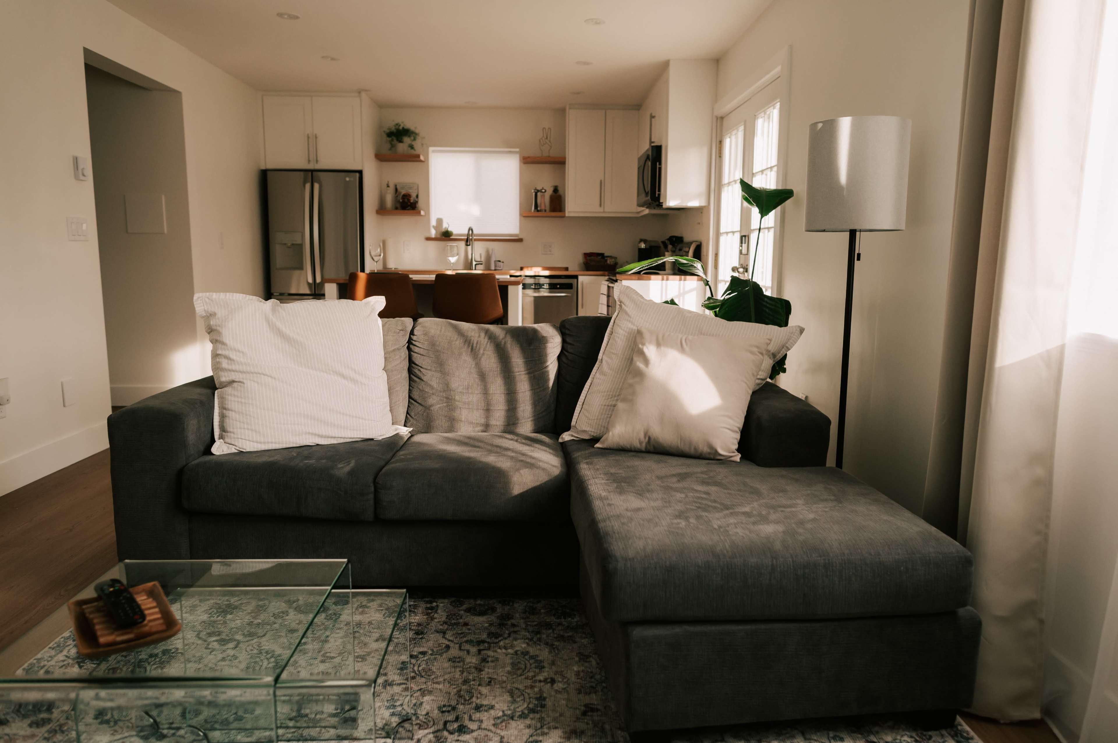 The image shows a modern living room with a grey sectional sofa, a glass coffee table, and an open kitchen visible in the background.