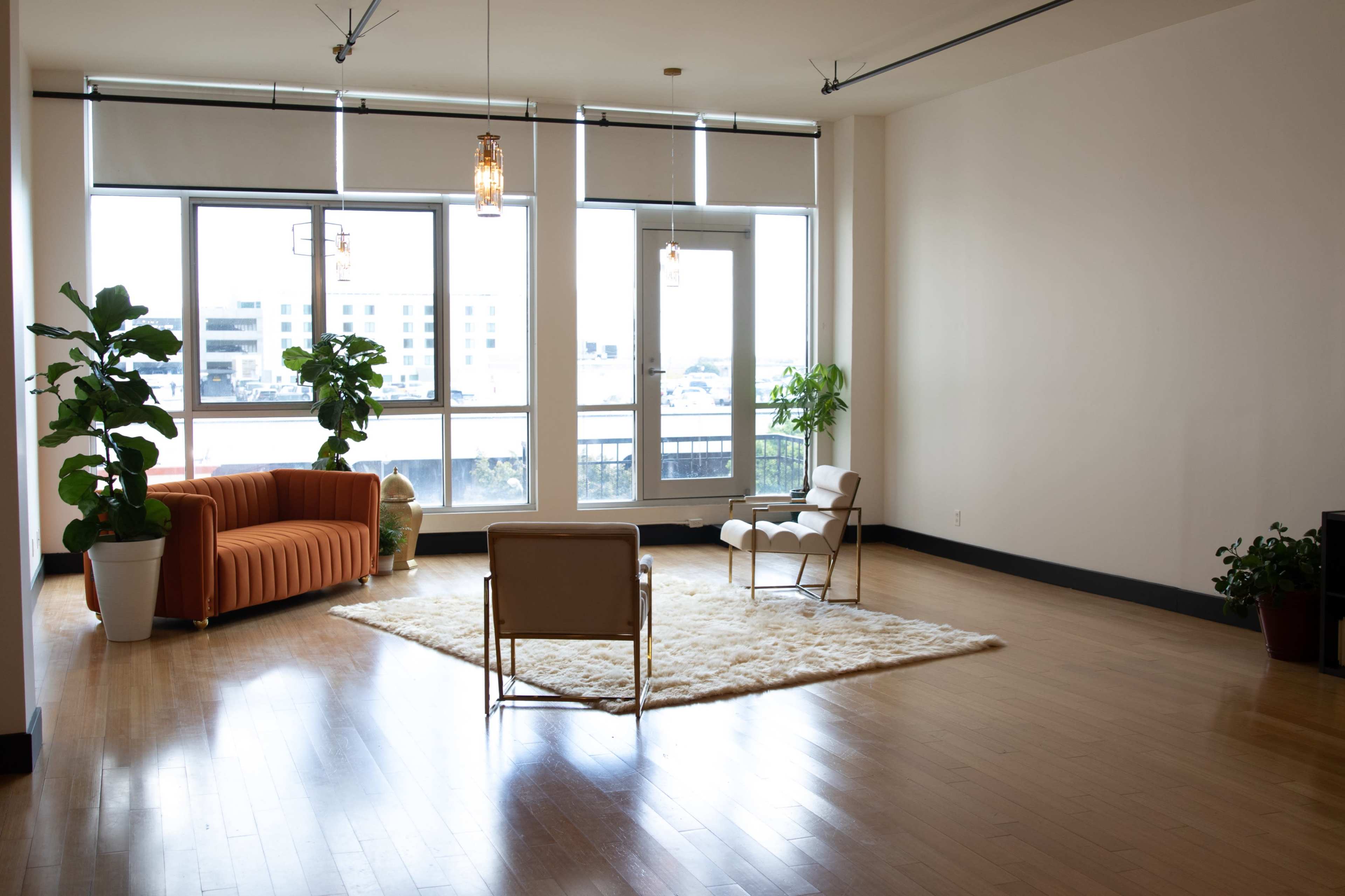A spacious, well-lit living room featuring an orange sofa, a white rug, and large windows with plants placed around the room.
