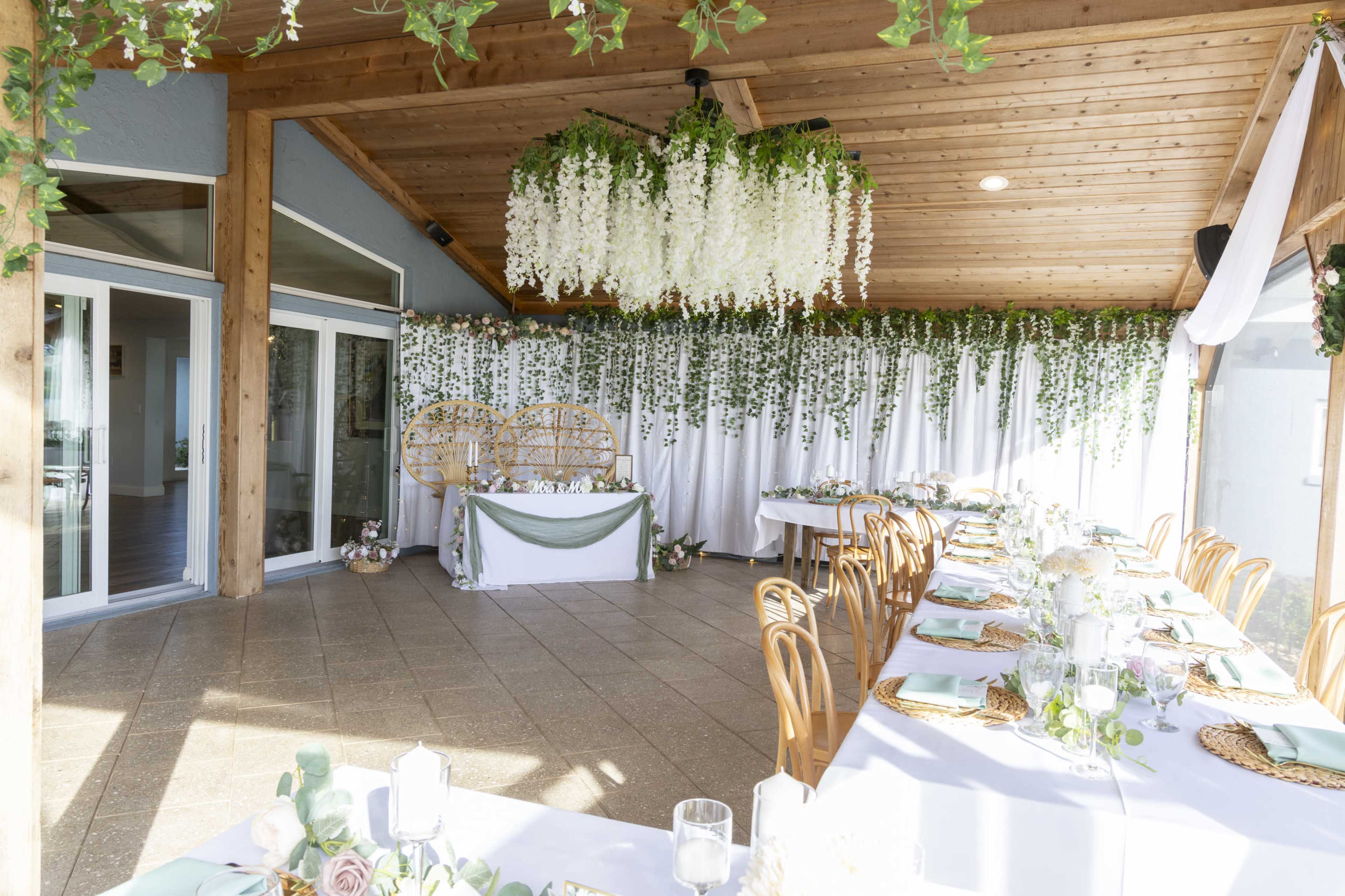 The image shows a beautifully arranged indoor event space with wooden beams, large windows, and tables set with floral centerpieces and elegant tableware.
