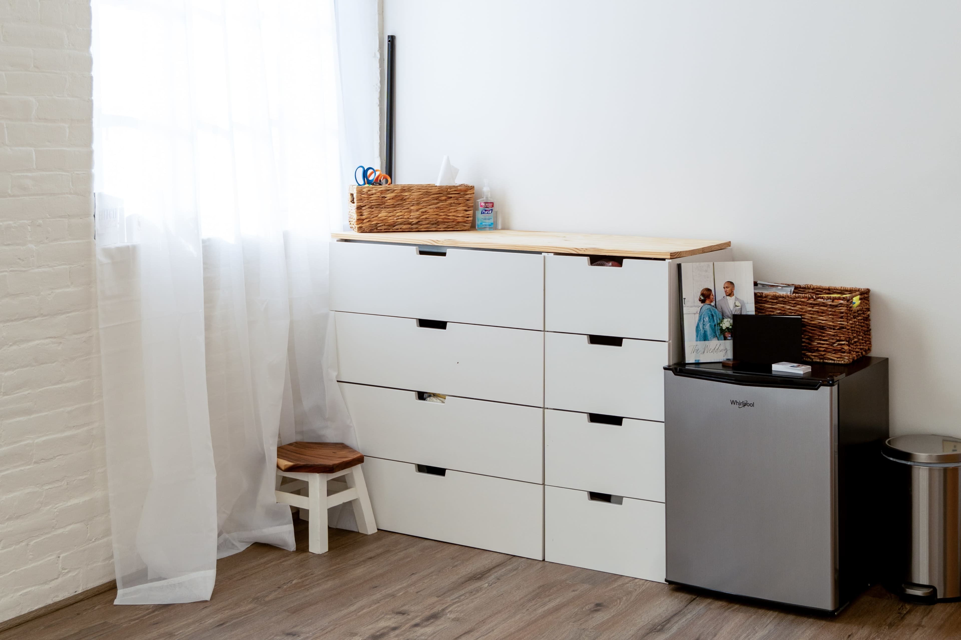 A simple interior scene features a white dresser with several drawers beside a small fridge and a chair, illuminated by natural light from a nearby window.