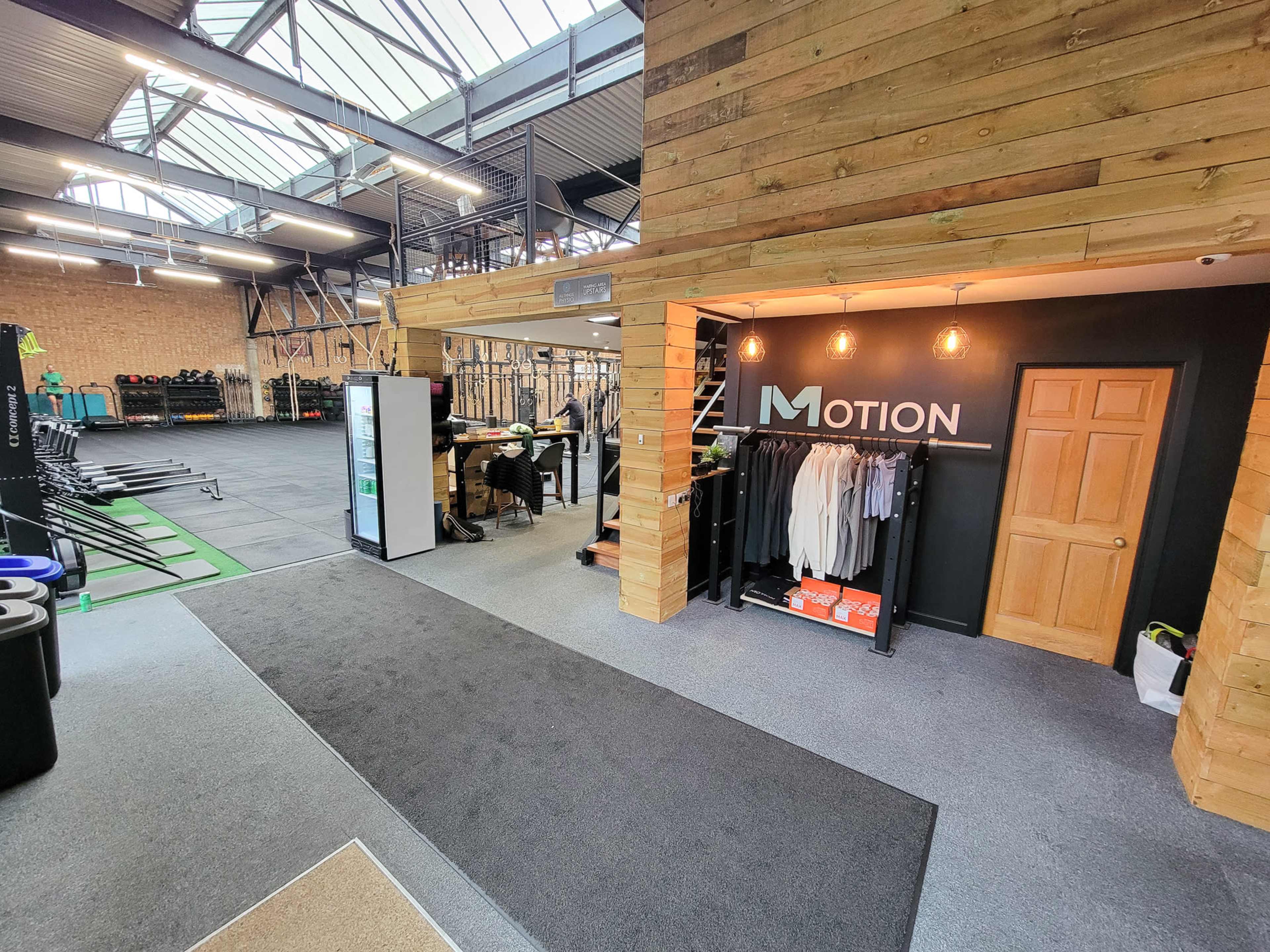 The image shows an interior view of a fitness facility featuring a reception area with wooden accents, workout equipment in the background, and a green exercise mat area.
