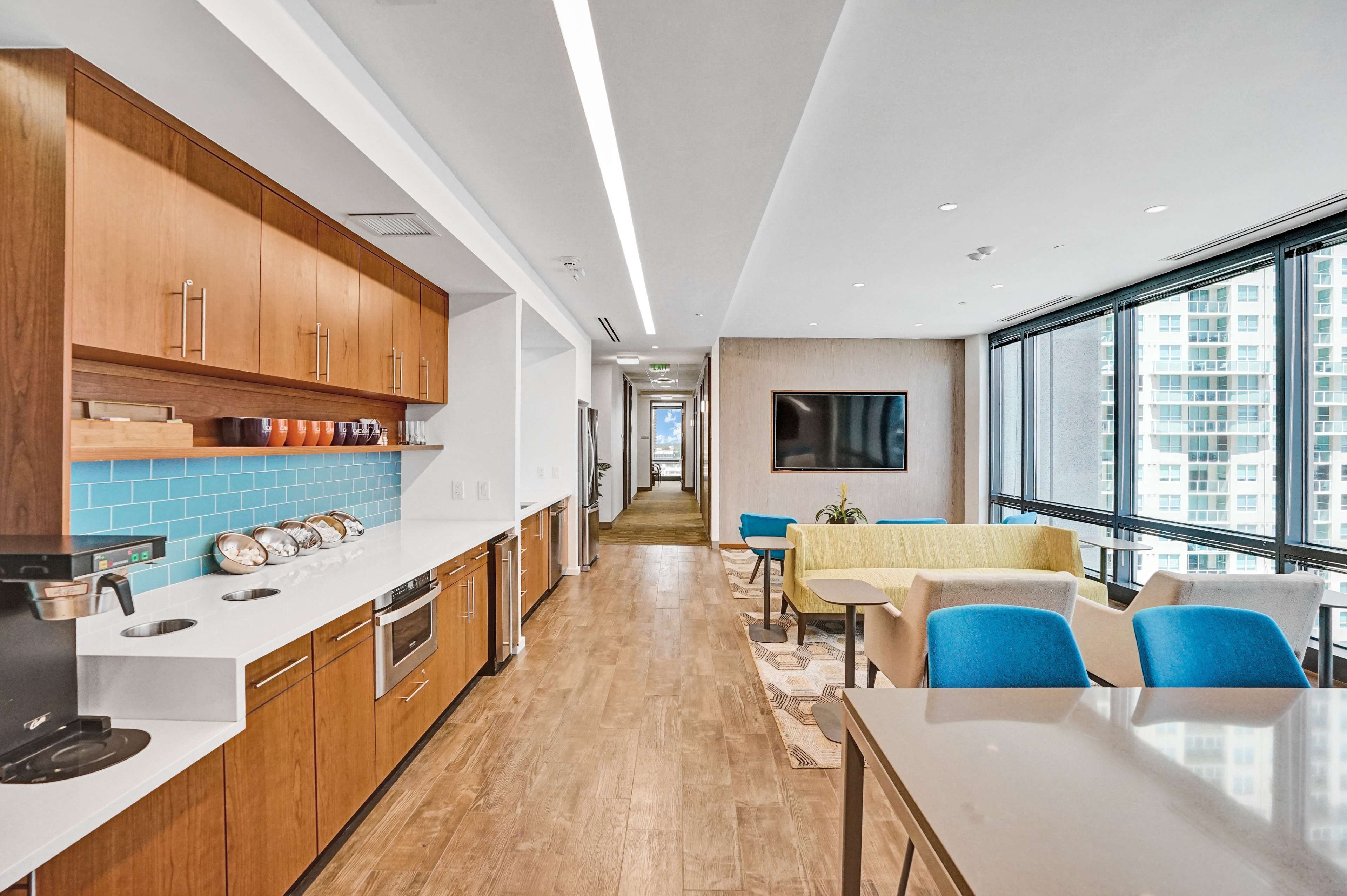 A sleek open-concept kitchen area features wooden cabinetry, a coffee machine, and a dining space with modern seating along large windows.