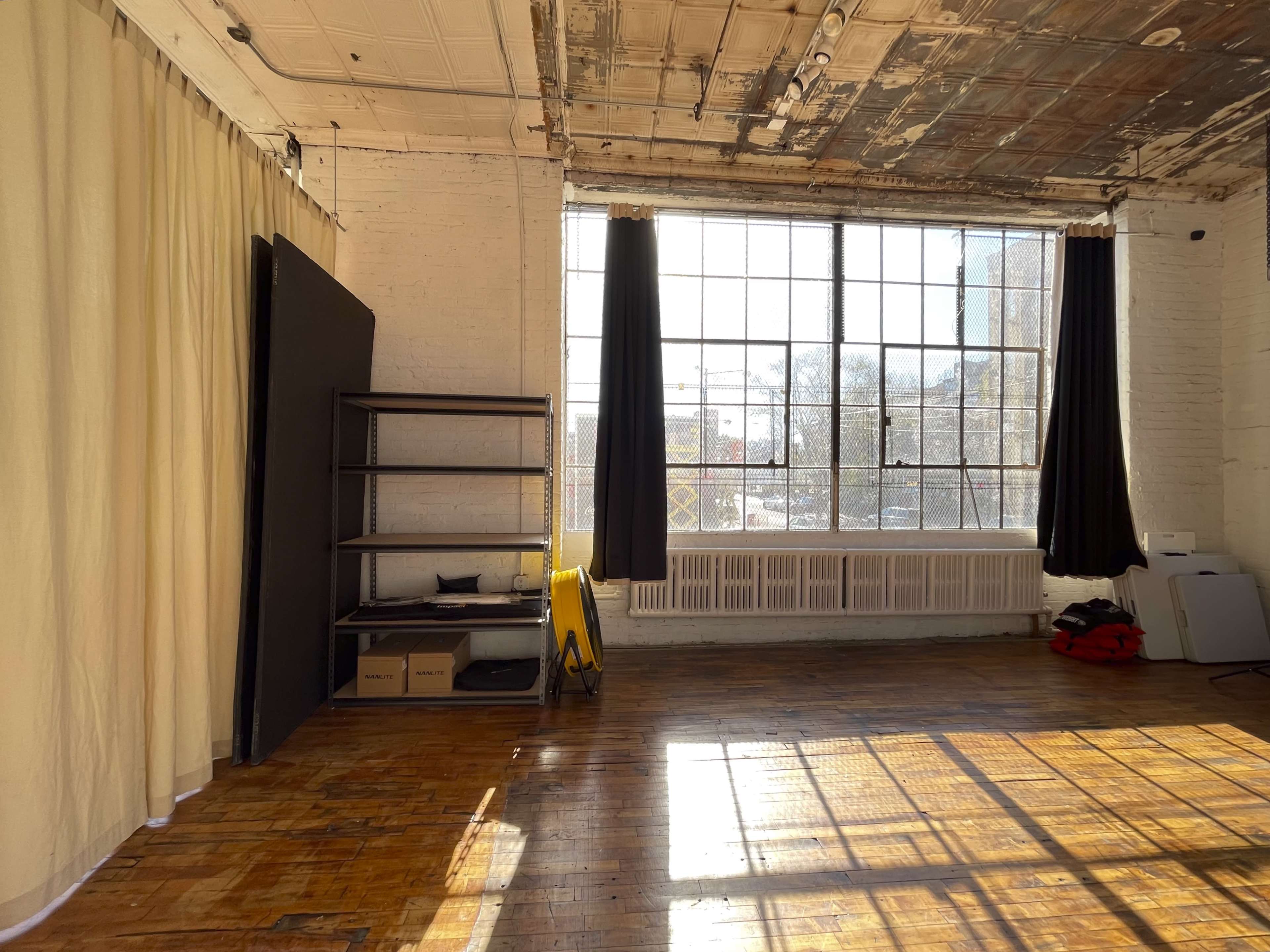 A spacious room with large windows, wooden floors, a shelf against the wall, and light filtering through black curtains.