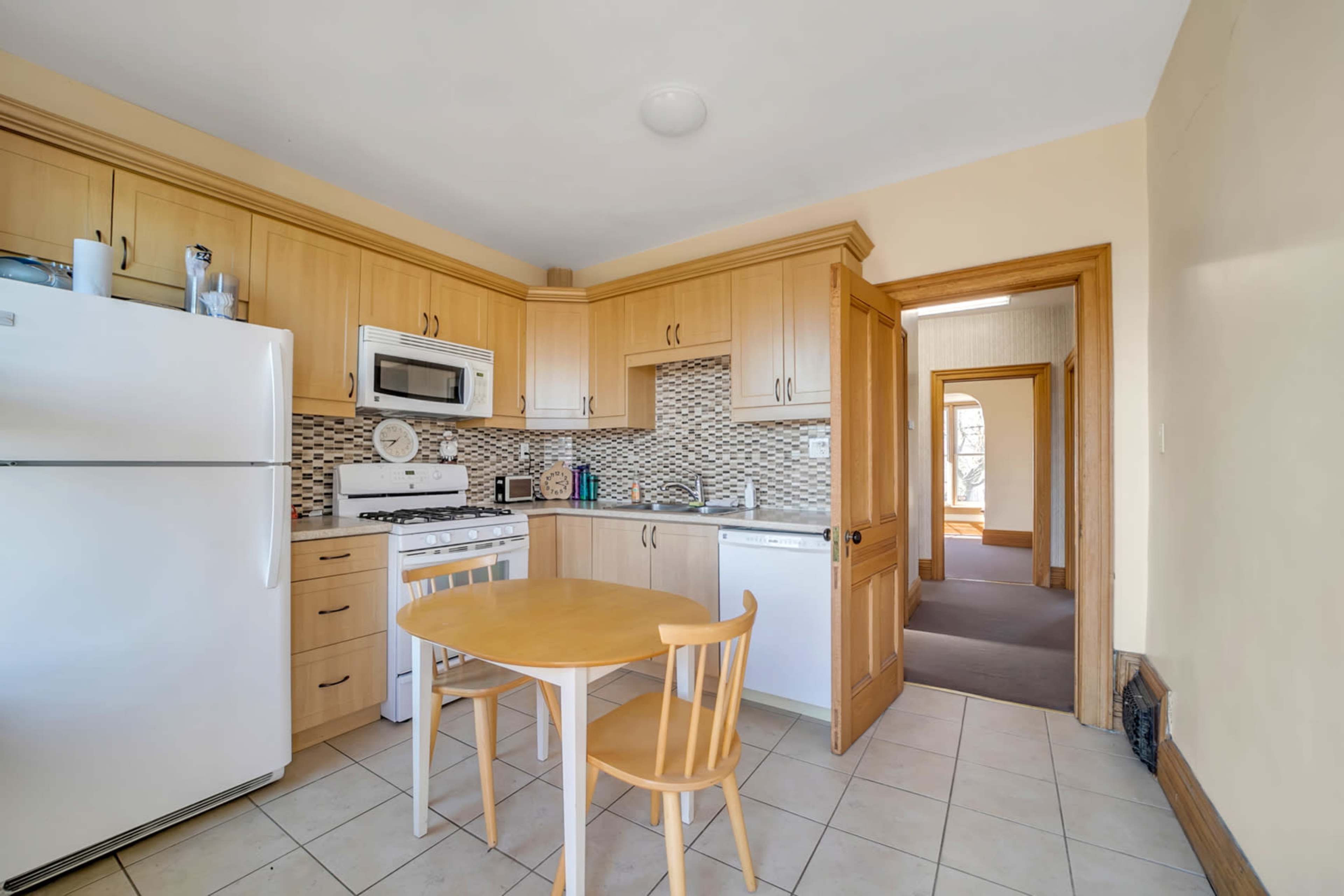 The image shows a small kitchen featuring wooden cabinets, a white refrigerator, a stove, and a table with two chairs, with a doorway leading to another room.