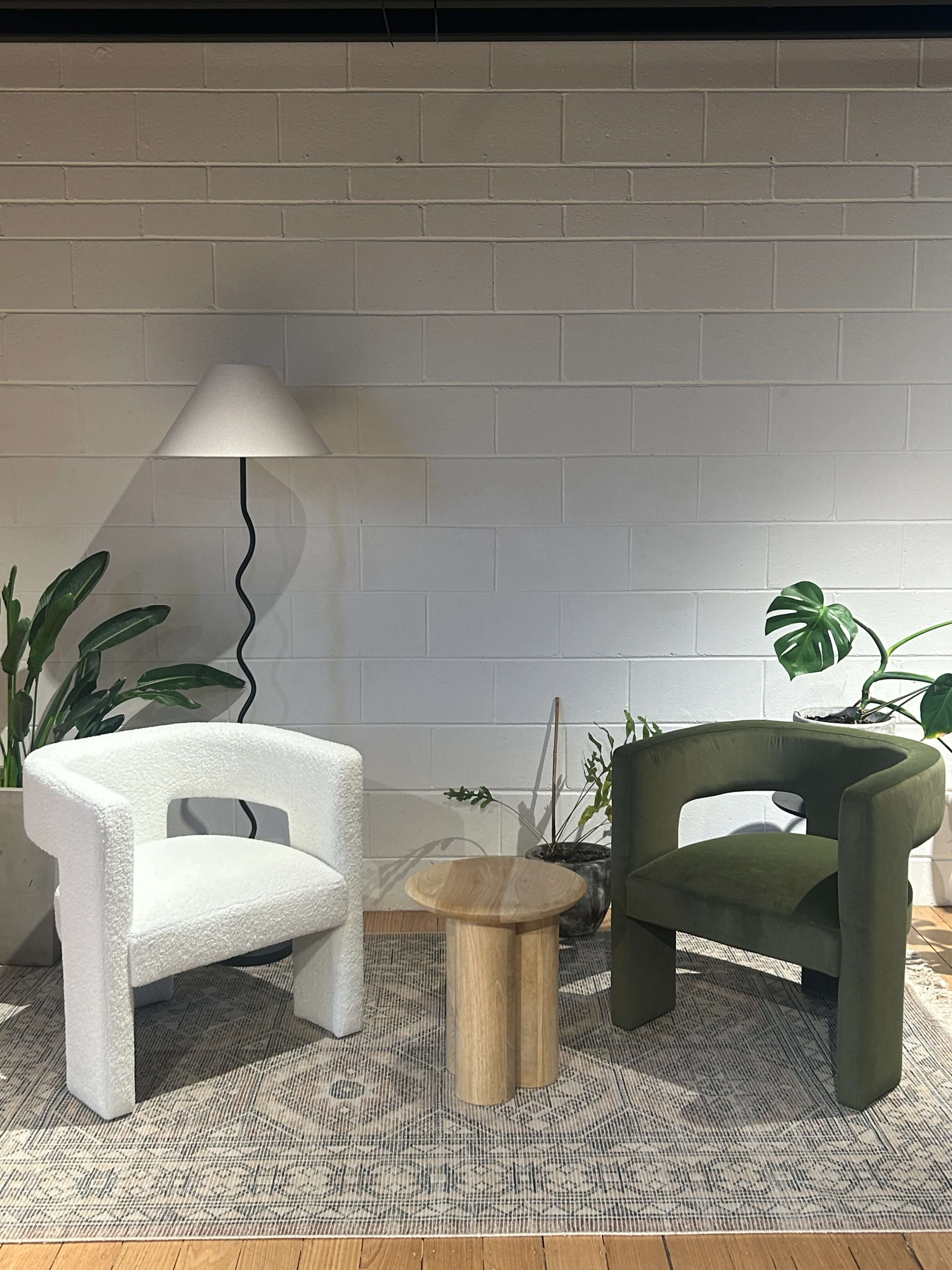 Two modern chairs, one white and one olive green, are positioned beside a round glass table on a patterned rug, with a tall lamp and potted plants in the background.