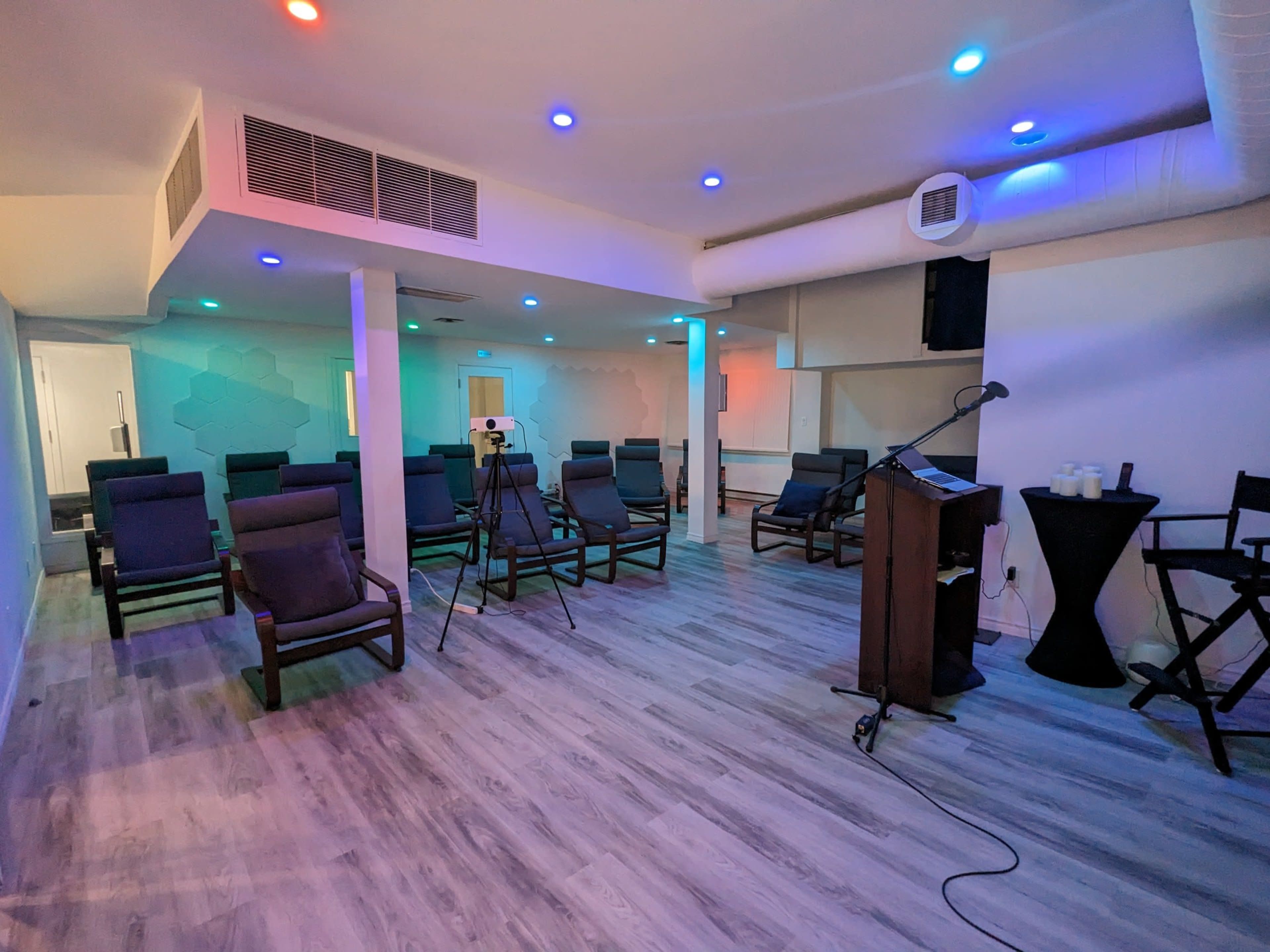 A spacious, well-lit room features rows of dark chairs, a podium, and a video camera set up for an event.